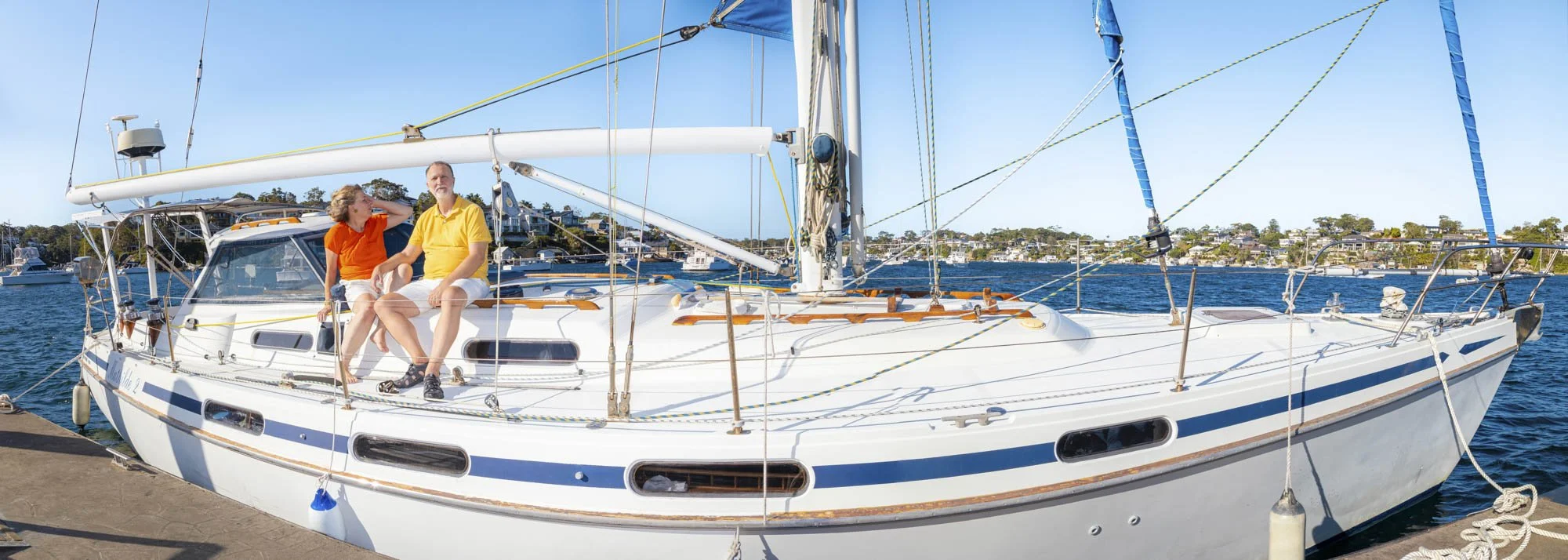 Two people sitting on a white sailboat docked at a marina, with a harbor and houses in the background on a sunny day.