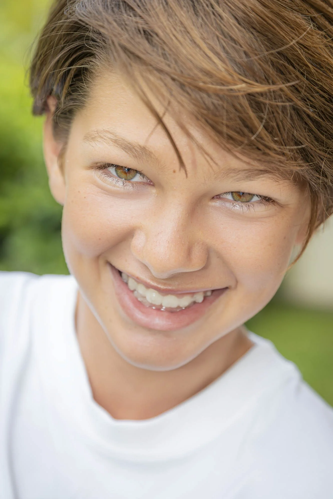 Close-up of a smiling young person with short reddish-brown hair and hazel eyes, wearing a white shirt, outdoors with blurred greenery in the background.