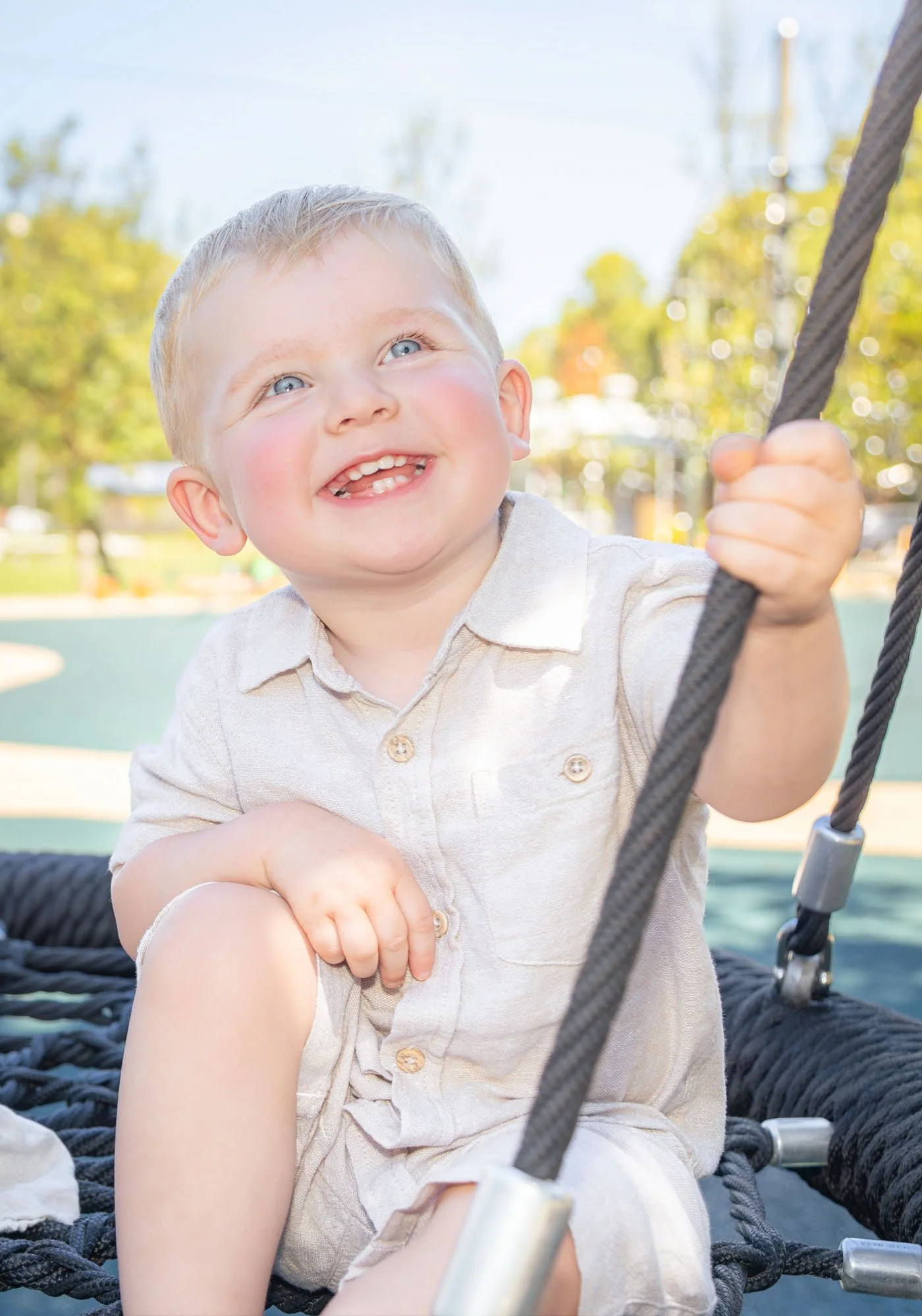 A young boy sitting on a playground structure with a bright smile, holding onto a black rope, outdoors in a park on a sunny day during a relaxed outdoor family photoshoot by Southern Exposure Photography.