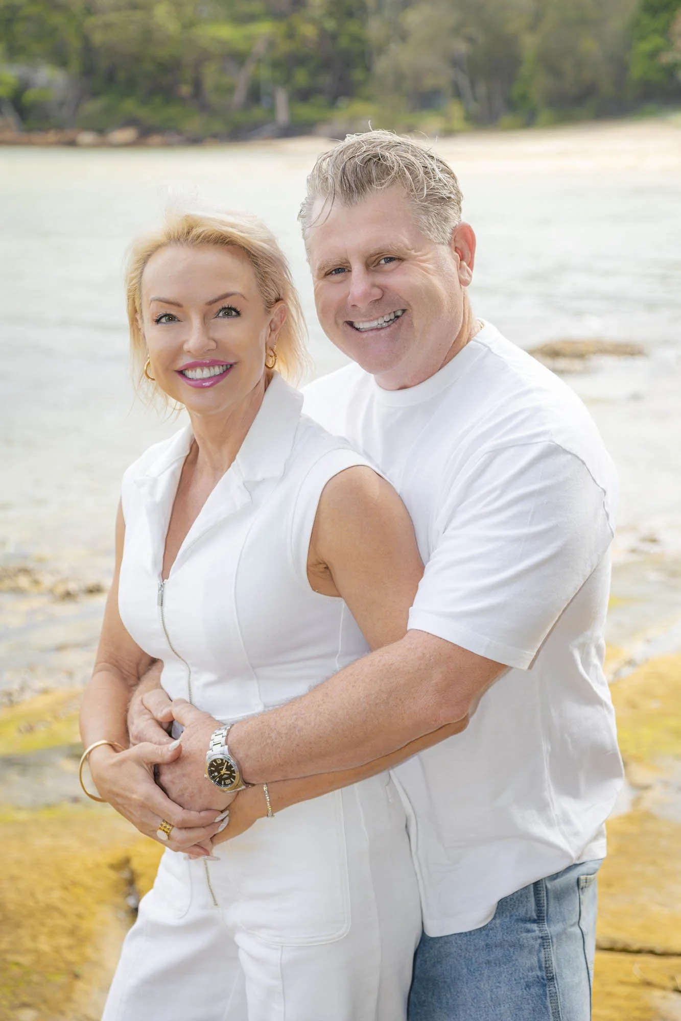 A smiling middle-aged couple in white clothing, embracing on a beach with water and trees in the background.