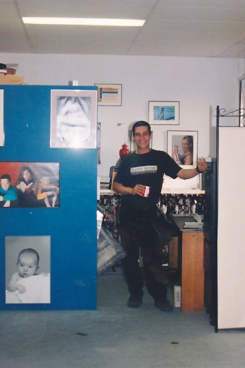 A man standing in an office, holding a mug and smiling at the camera. There are pictures on a blue partition and framed photos on the wall behind the person in a studio in Cronulla.