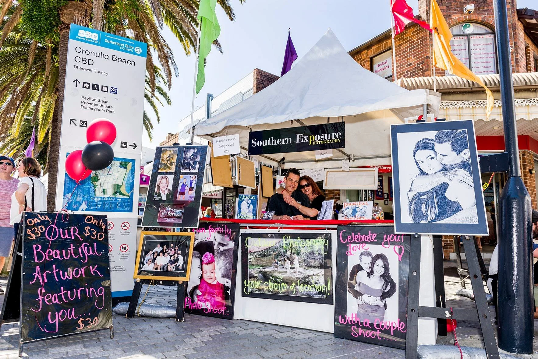 A photography booth at a market in Cronulla with various framed photographs, with a large blackboard sign advertising photography artwork for $30, and a vendor couple sitting inside the tent, surrounded by palm trees and people walking by.