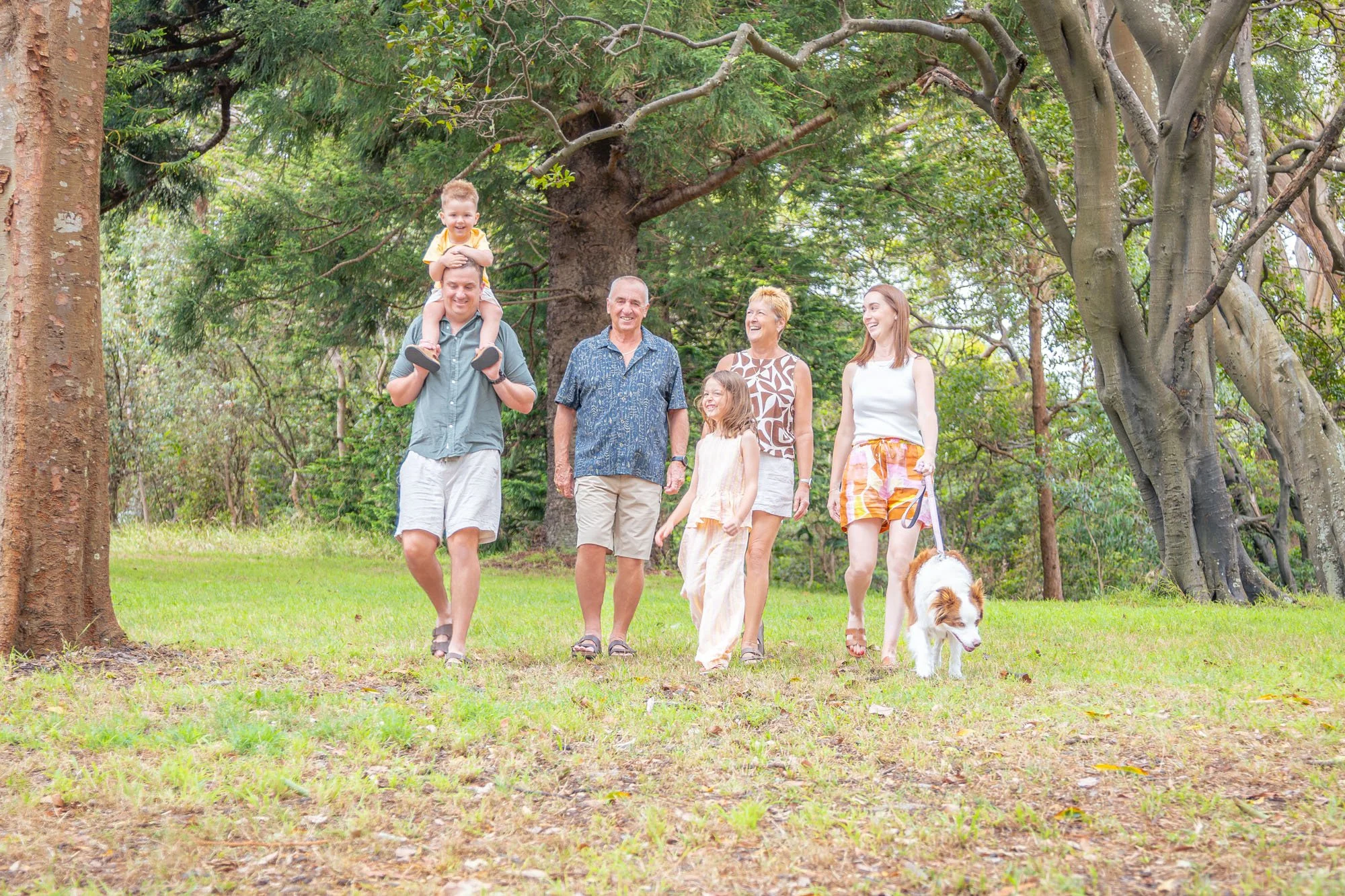 A multigenerational family of six with a dog walking in a park with lush green trees and grass. They are smiling and enjoying a sunny day during a relaxed outdoor family photoshoot by Southern Exposure Photography.