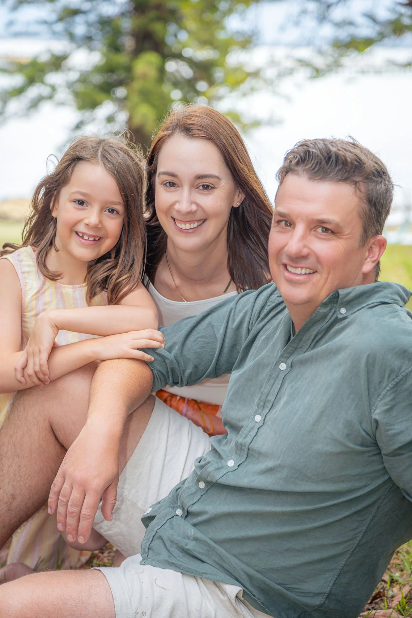 A smiling family of three sitting outdoors on grass, with trees in the background. The young girl has long brown hair, the woman has straight brown hair, and the man has short curly hair. They are all close together, enjoying a sunny day during a rel