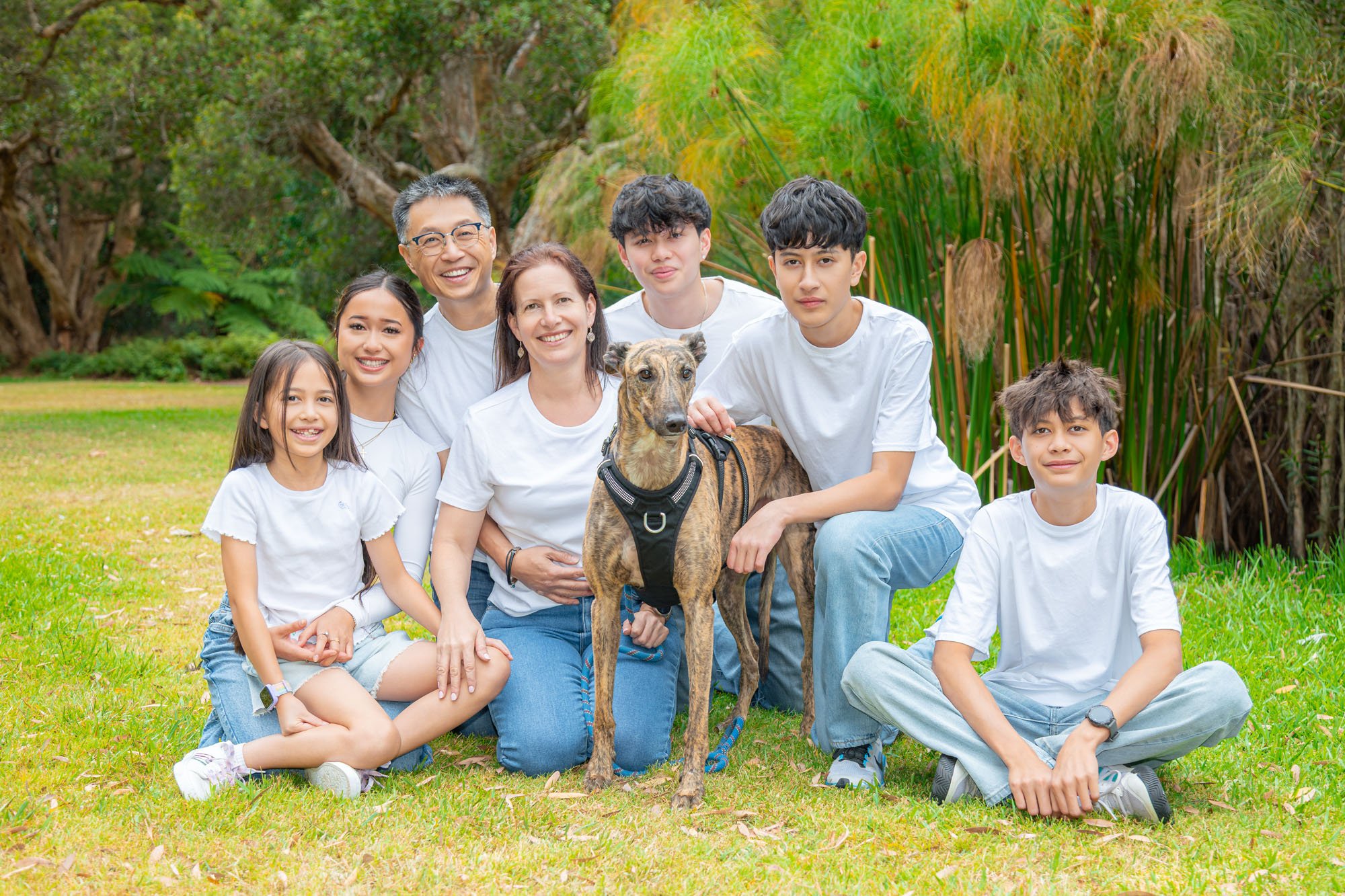 A large family groupof seven people and a dog posing outdoors in a lush green park during a relaxed outdoor family photoshoot by Southern Exposure Photography.