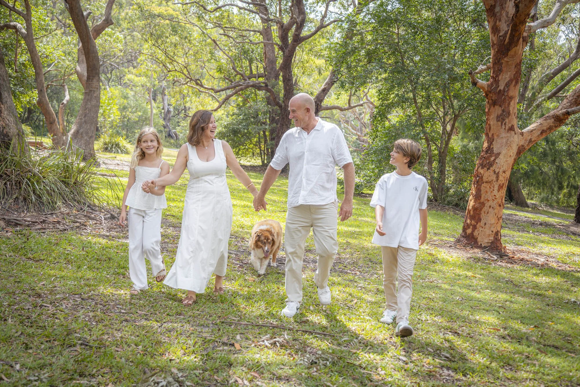 A family of five and a dog walking through a sunlit park with tall trees, all dressed in white and beige, smiling and holding hands.