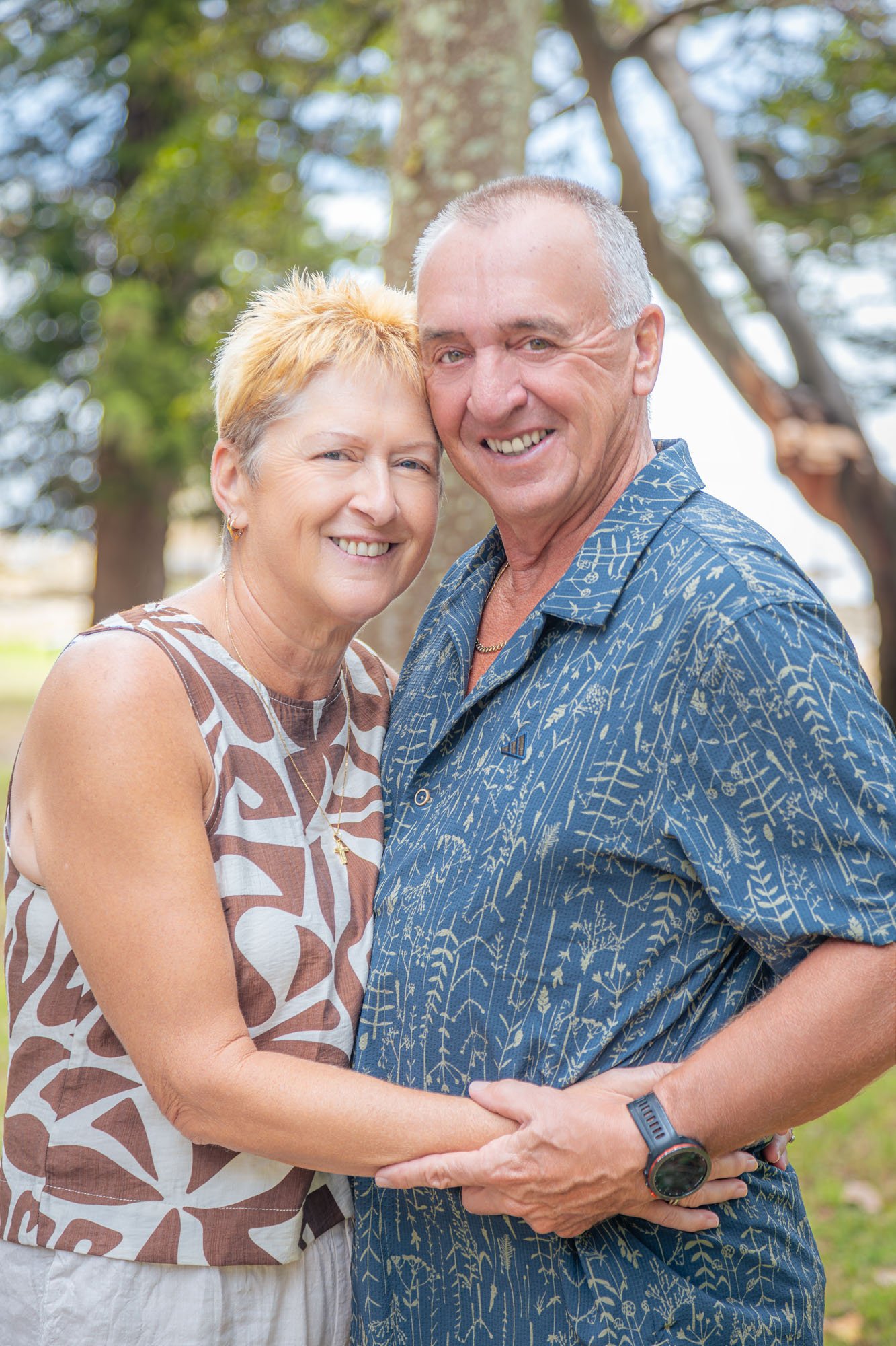 A smiling senior couple stands outdoors, holding hands and embracing each other with trees in the background during a relaxed outdoor family photoshoot by Southern Exposure Photography.