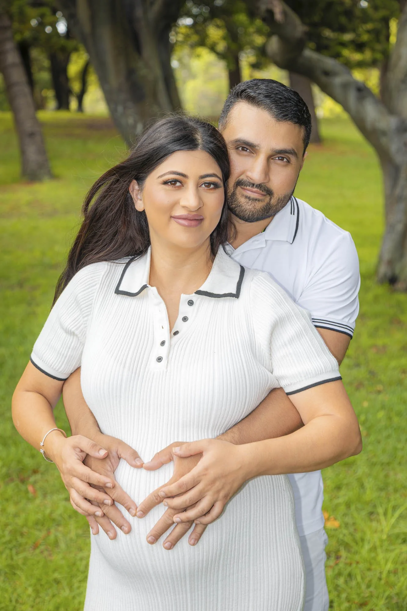 A smiling pregnant woman in a white shirt making a heart shape with her hands on her belly, with a man hugging her from behind in a park.