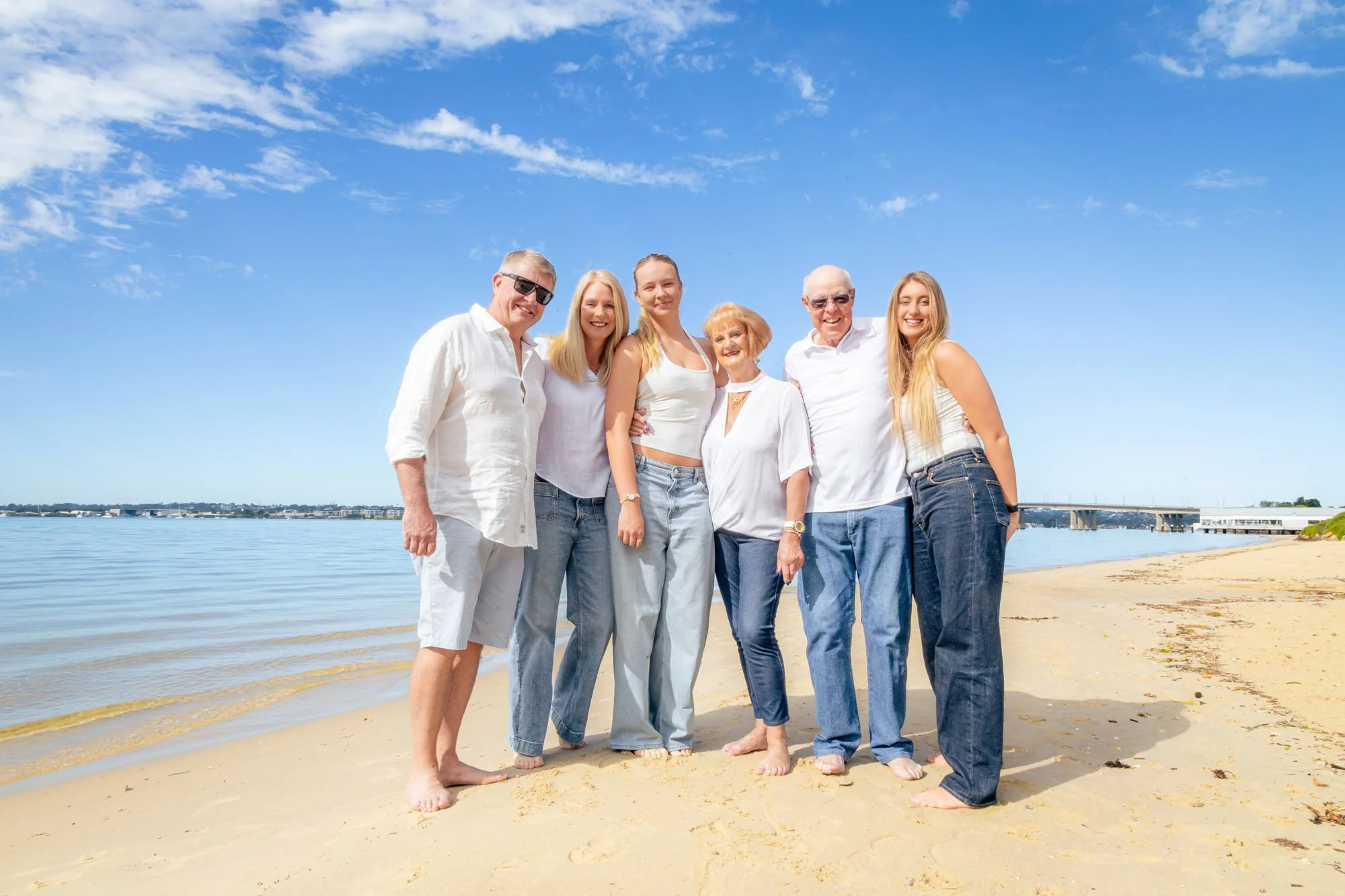 A group of six people standing together on a beach under a blue sky with some clouds during a relaxed outdoor family photoshoot by Southern Exposure Photography.