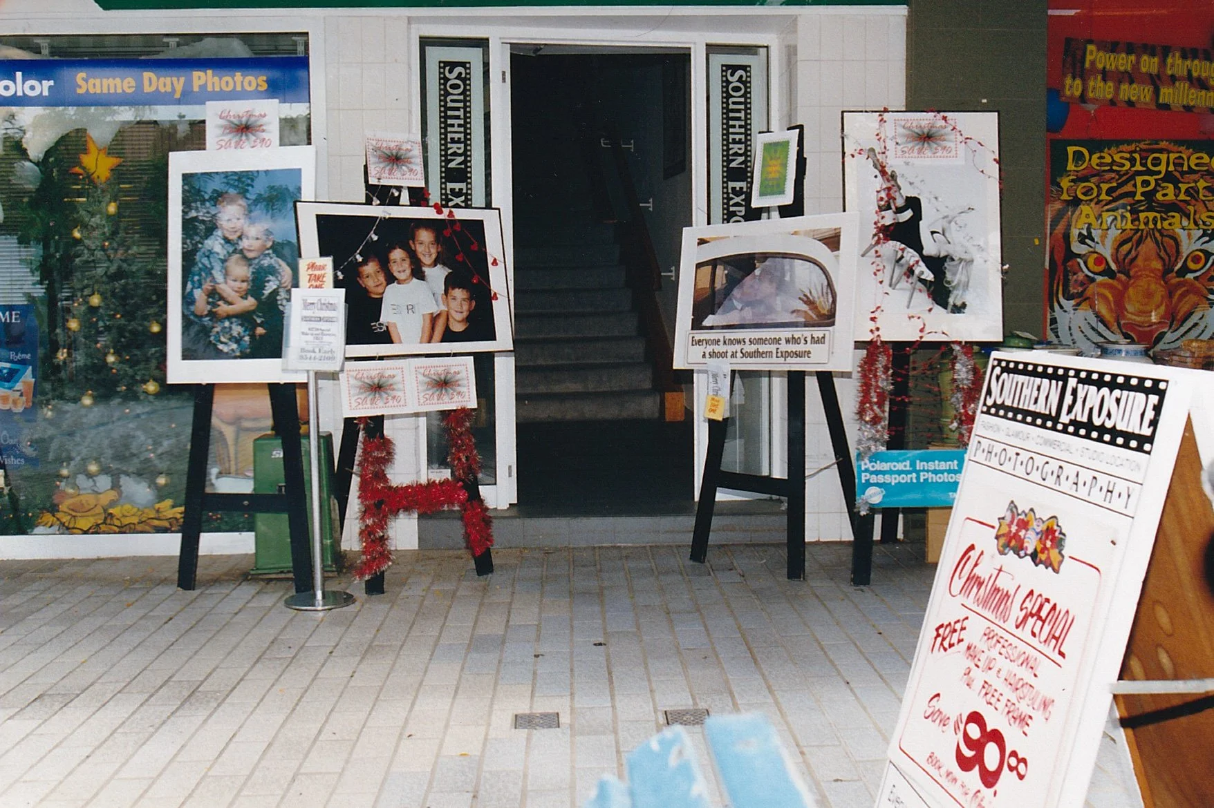 A photo of a storefront with photo frames, easles and Christmas decorations and several signs and posters promoting Southern Exposure Photography's photography services, including instant passport photos and special Christmas offers.