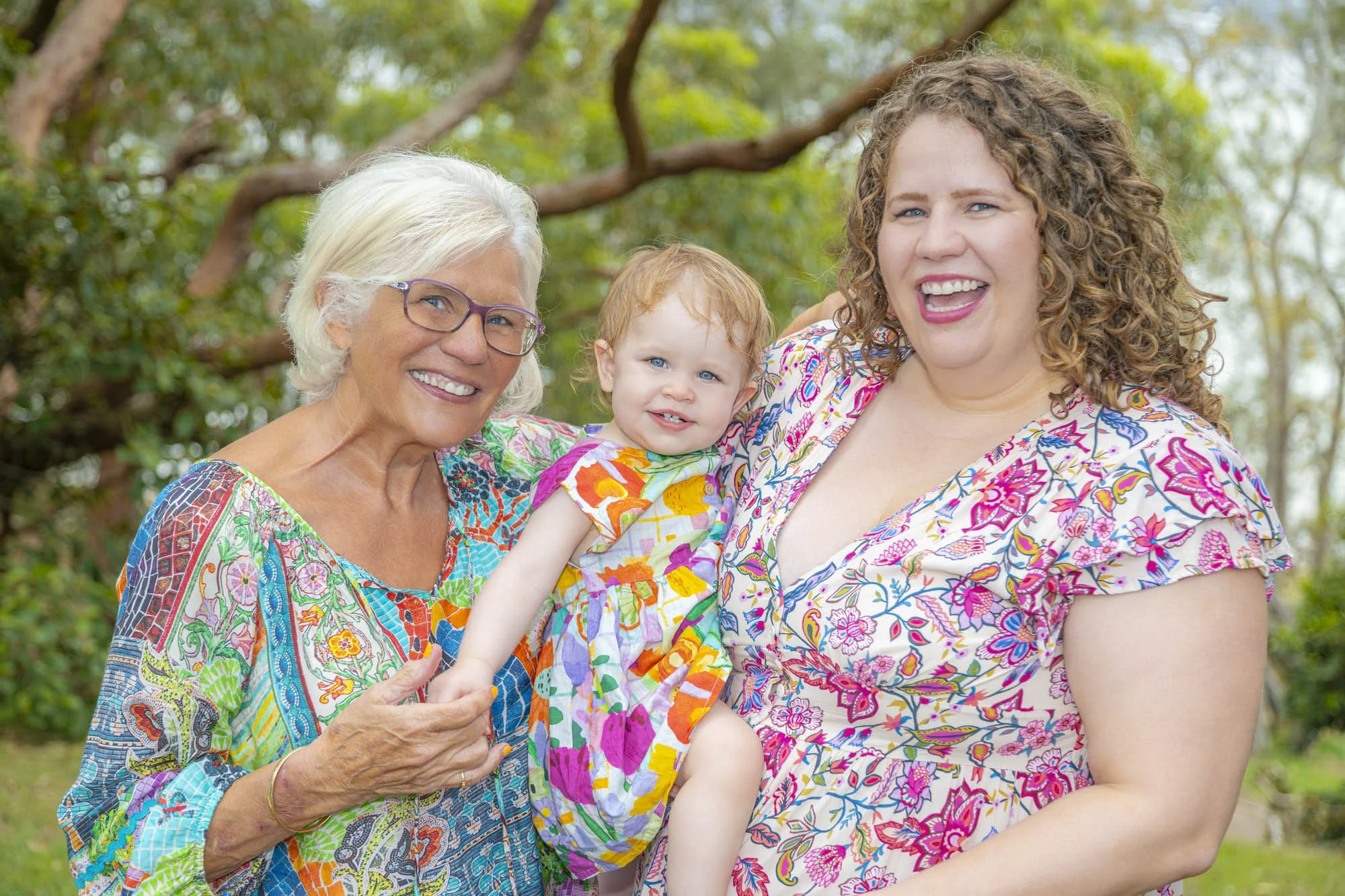 Three generations of women smiling outdoors. An elderly woman with glasses, a middle-aged woman, and a young girl in a colorful dress. They are in a garden with trees in the background.