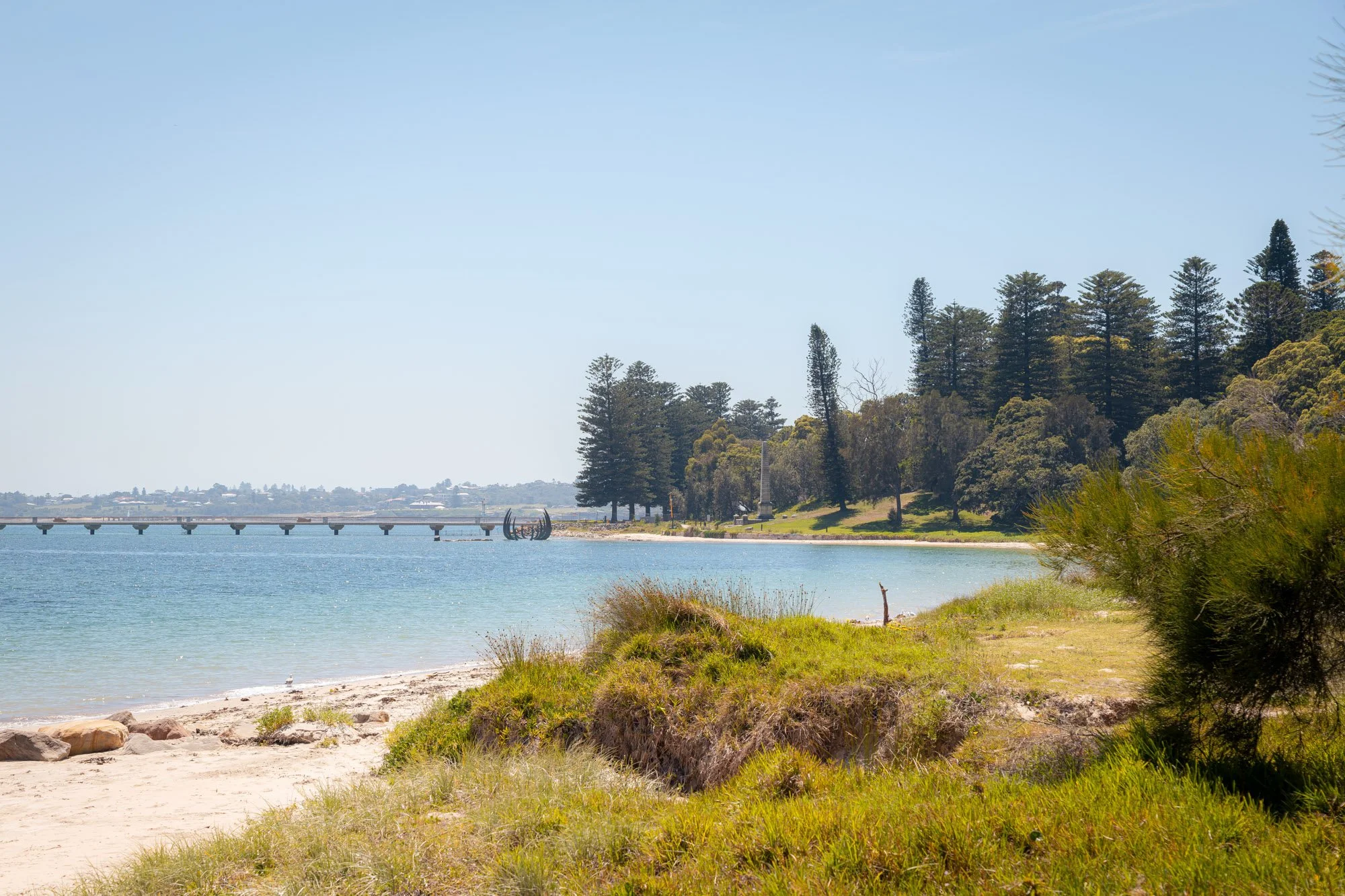 An image of Kamay Botany Bay National Park. It features a sandy beach with green grass and bushes, calm water, trees on the shoreline, and a pier extending into the water under a clear blue sky.