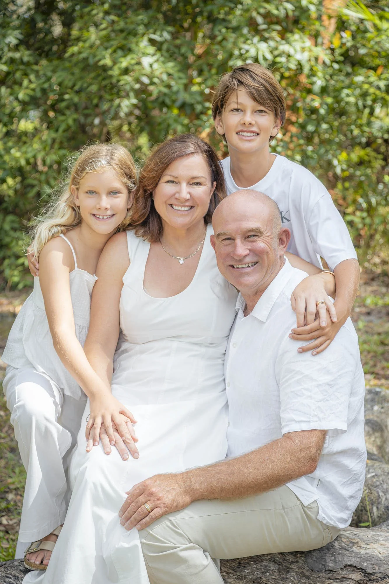 A happy family of four outdoor with green trees, the mother, father, daughter, and son all wearing white, smiling and sitting together.