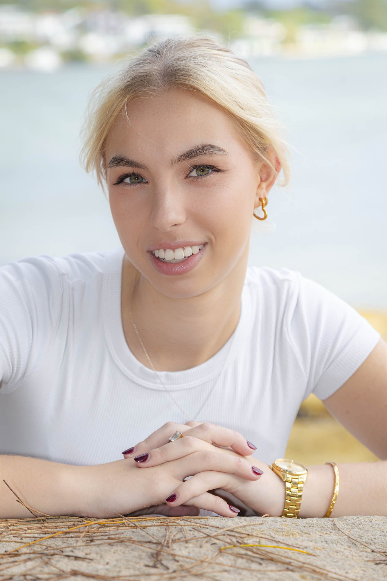 A young woman with short blonde hair, wearing a white t-shirt, jewelry, and a watch, is smiling while leaning on a wooden surface outdoors with water and trees in the background.