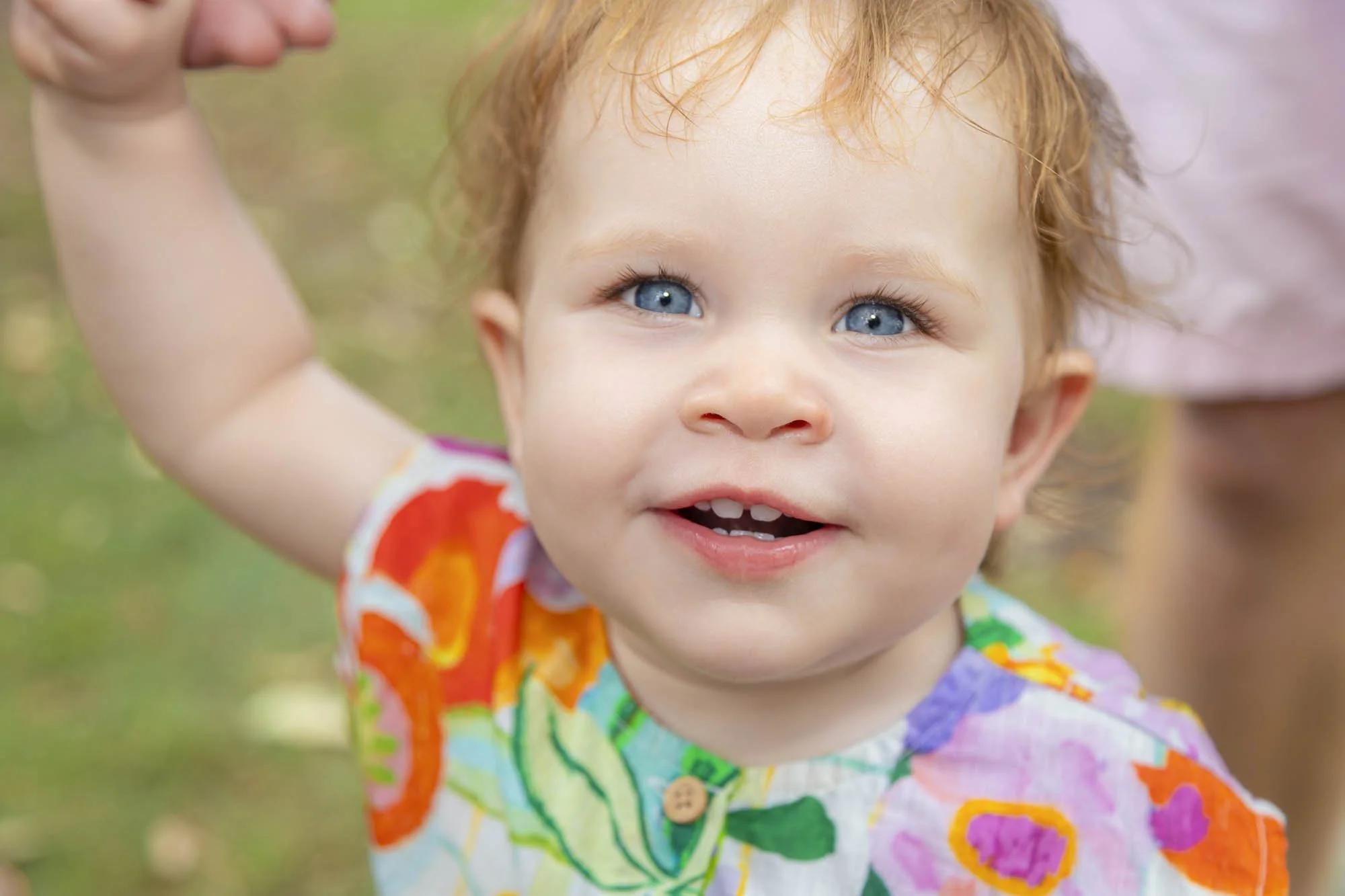 Close-up of a smiling toddler with blue eyes and red hair, wearing a colorful shirt, outdoors.