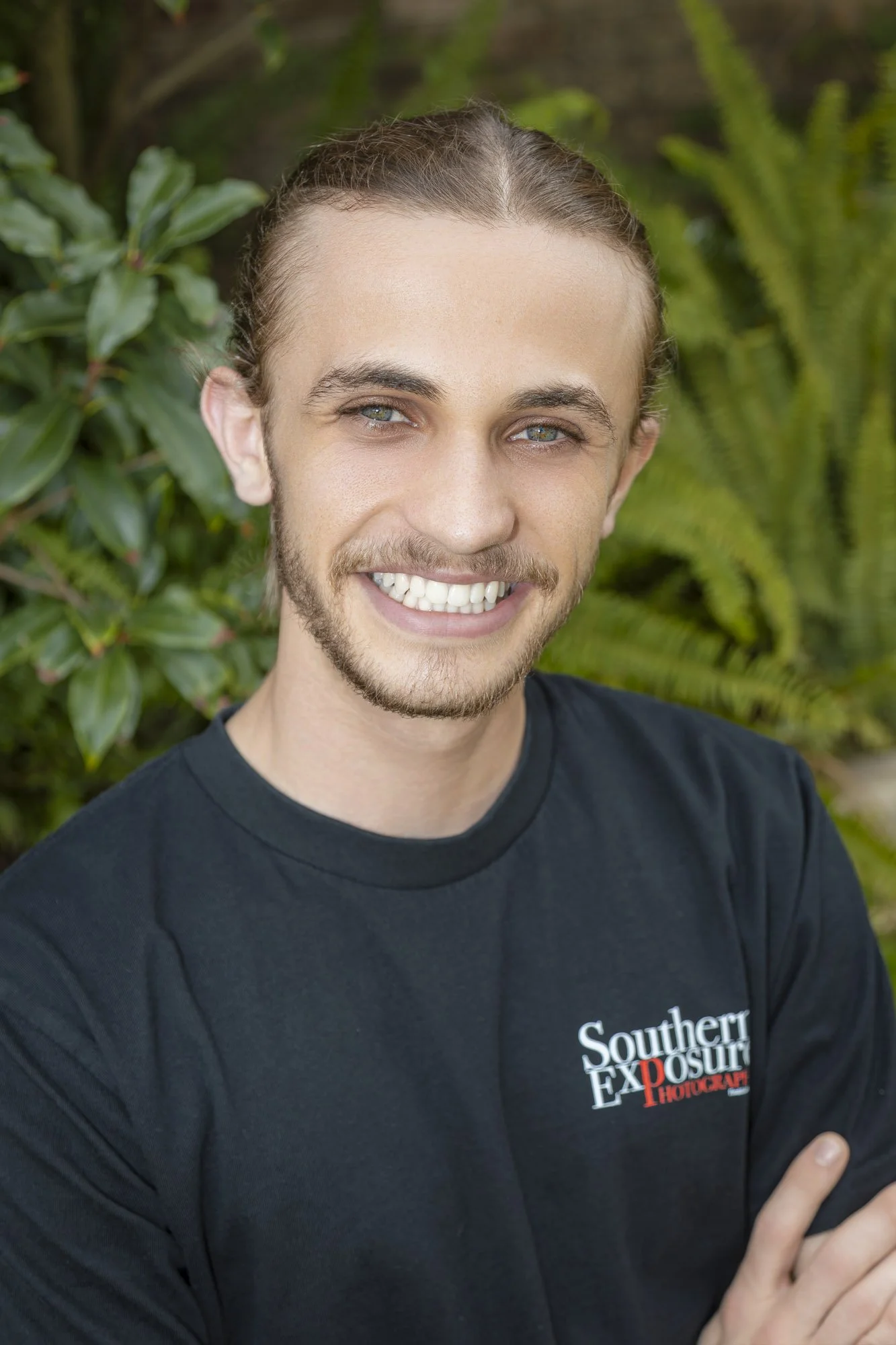A young man with long hair, a beard, and blue eyes smiling outdoors, wearing a black t-shirt with 'Southern Exposure Photography' logo, with greenery background.