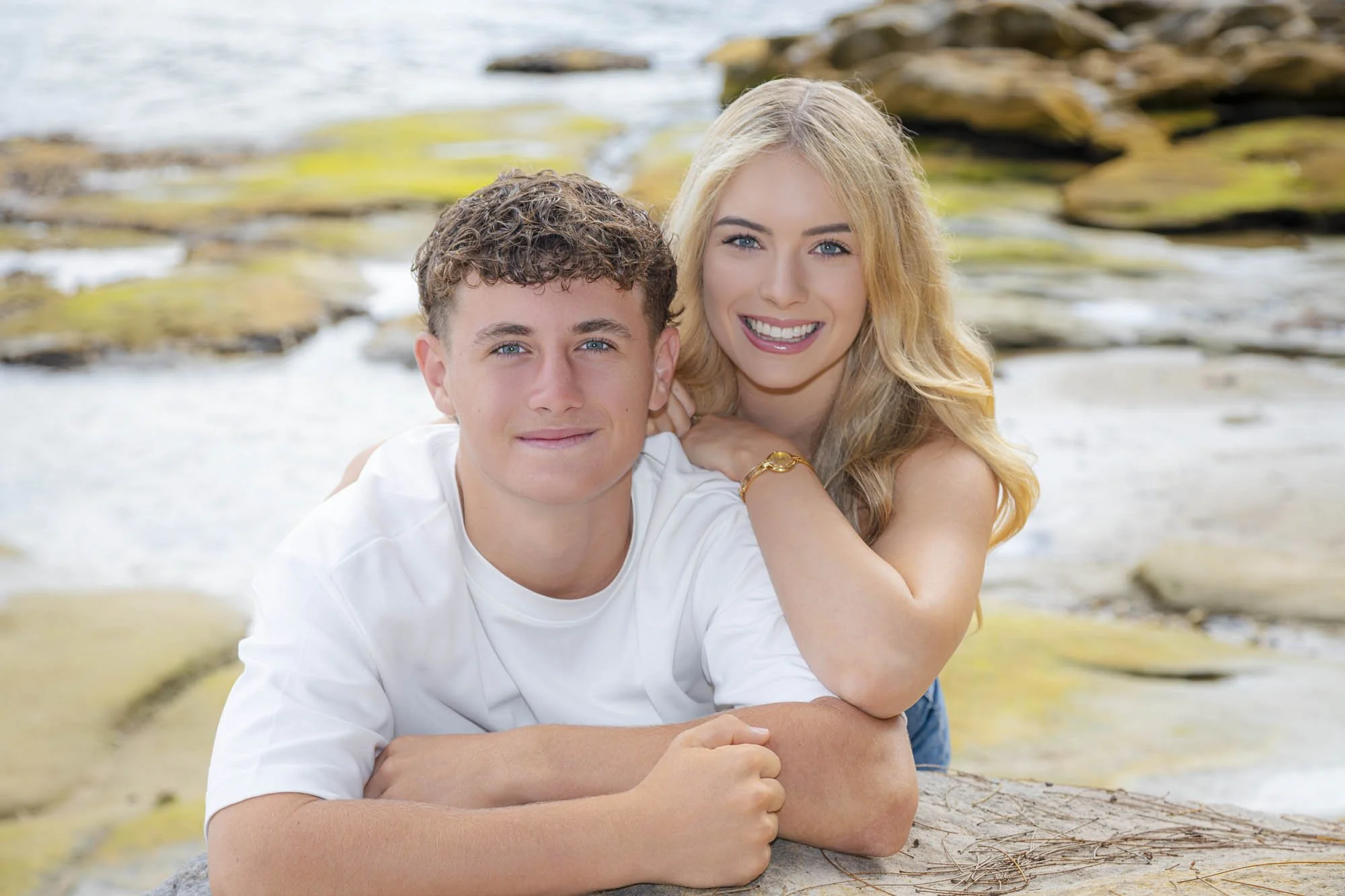 A young man and woman lying on a rock by the water, smiling at the camera with rocks and water in the background.