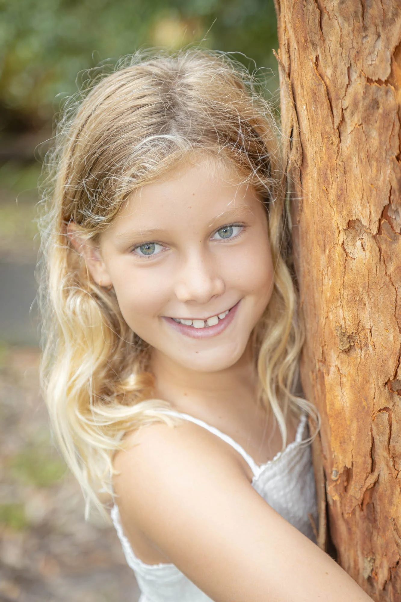 A young girl with blonde wavy hair and blue eyes smiling while hugging a tree outdoors.