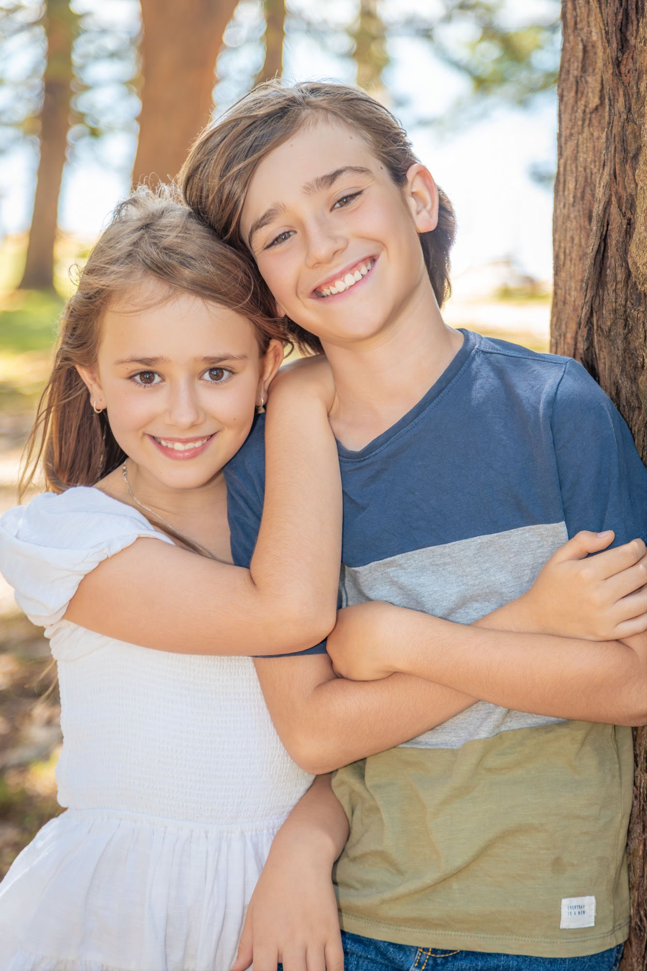 A brother and sister smiling, standing outdoors near a tree, with arms around each other during a relaxed outdoor family photoshoot by Southern Exposure Photography.