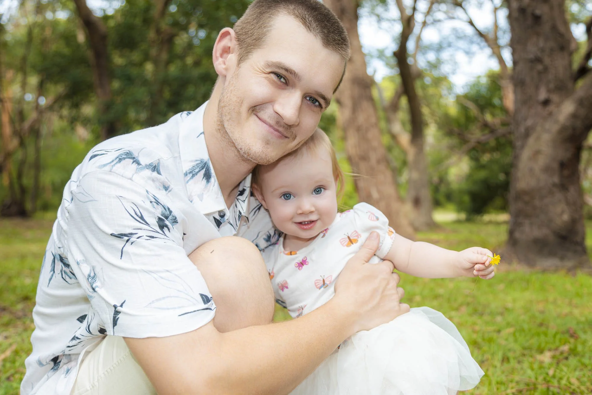 A man and a young girl outdoors in a park with trees in the background, smiling and holding a yellow flower.