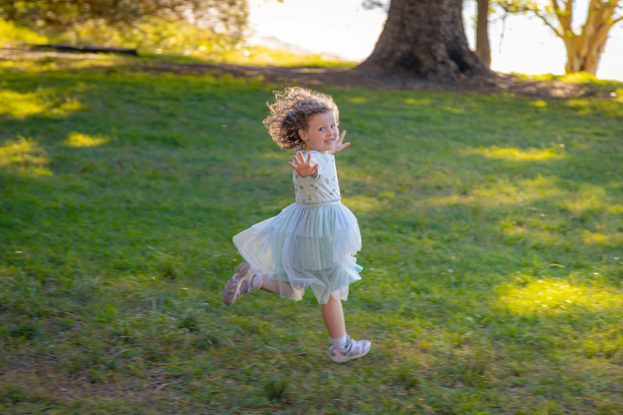A young girl with curly hair running on grass in a park, wearing a light blue dress and pink sneakers, with sunlight filtering through trees in the background during a relaxed outdoor family photoshoot by Southern Exposure Photography.