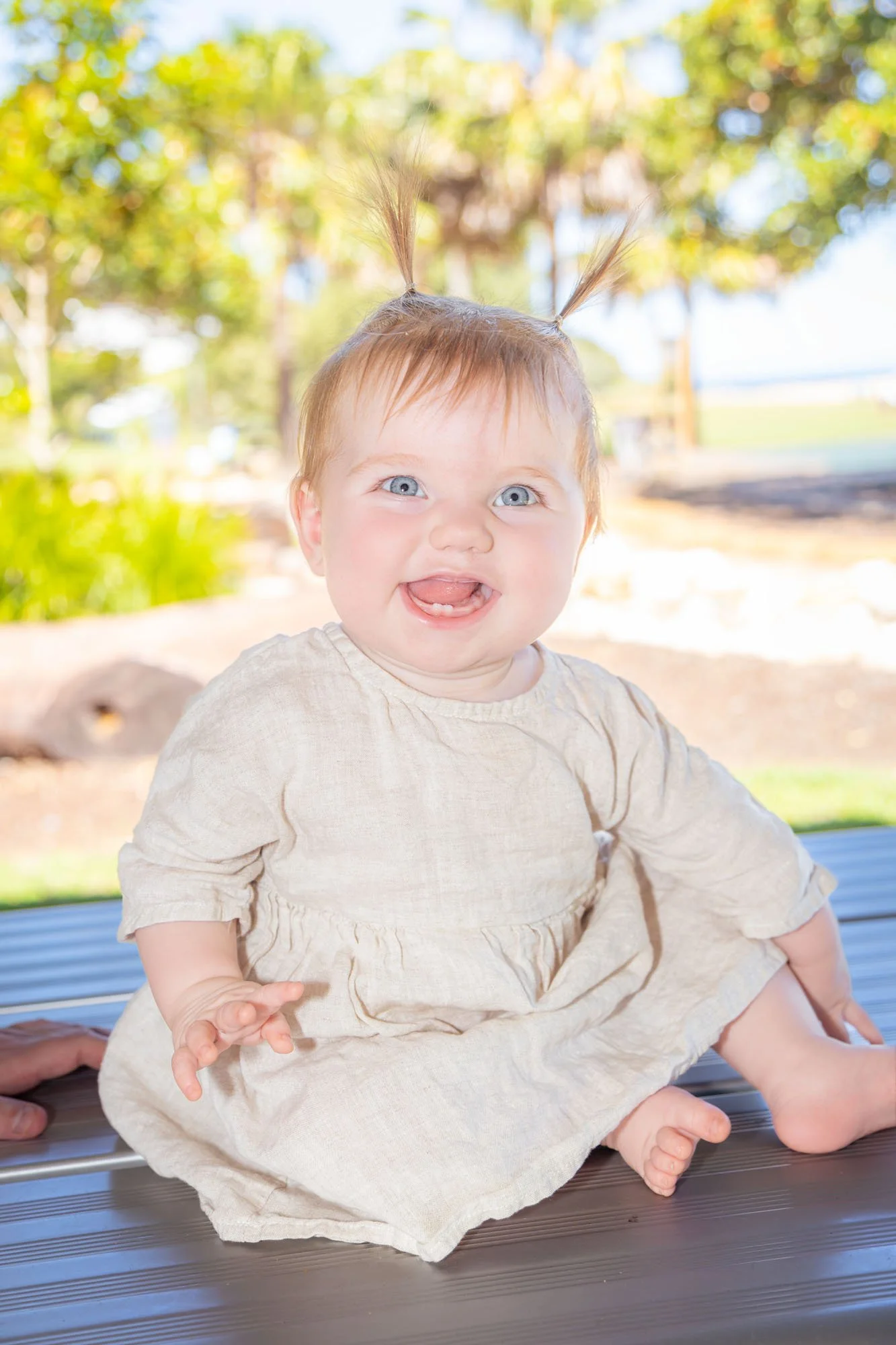 A smiling baby girl with blue eyes and sticking out tongue, sitting outdoors on a bench, with trees and greenery in the background during a relaxed outdoor family photoshoot by Southern Exposure Photography.
