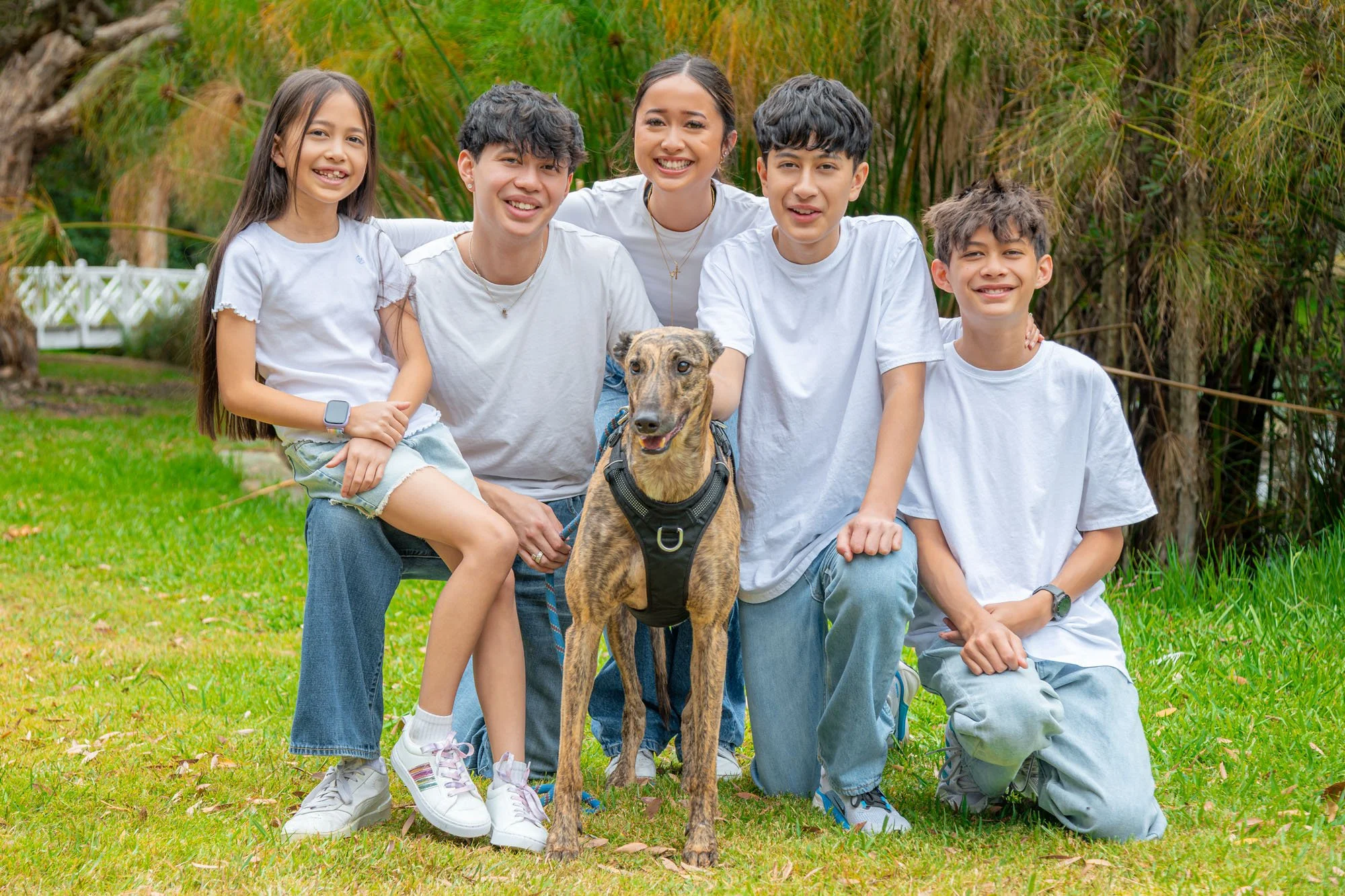 A group of five kids and a dog smiling outdoors on green grass with trees in the background during a relaxed outdoor family photoshoot by Southern Exposure Photography.