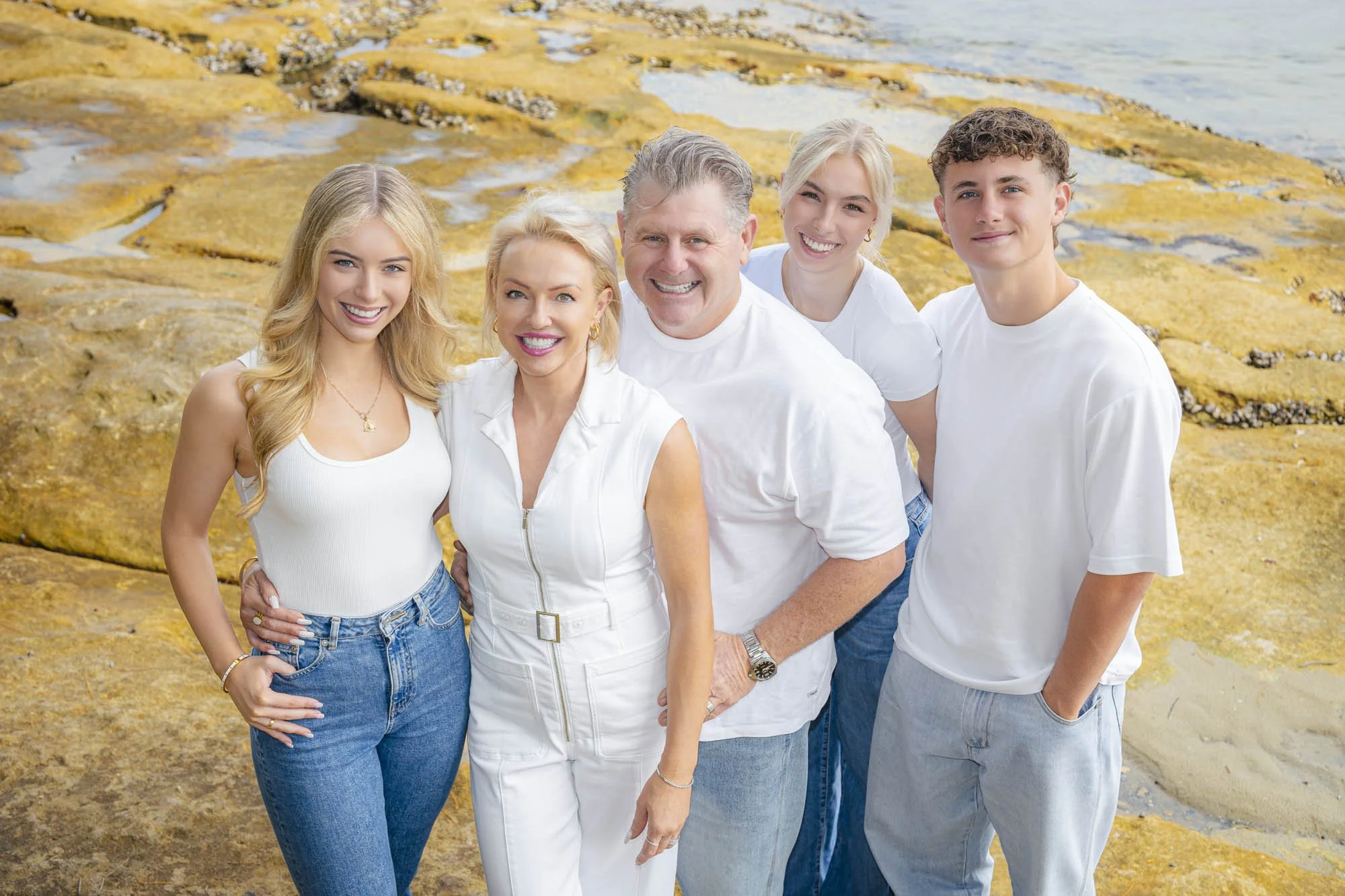A happy family of five standing on a rocky beach near the water, all smiling and dressed in white and casual jeans.