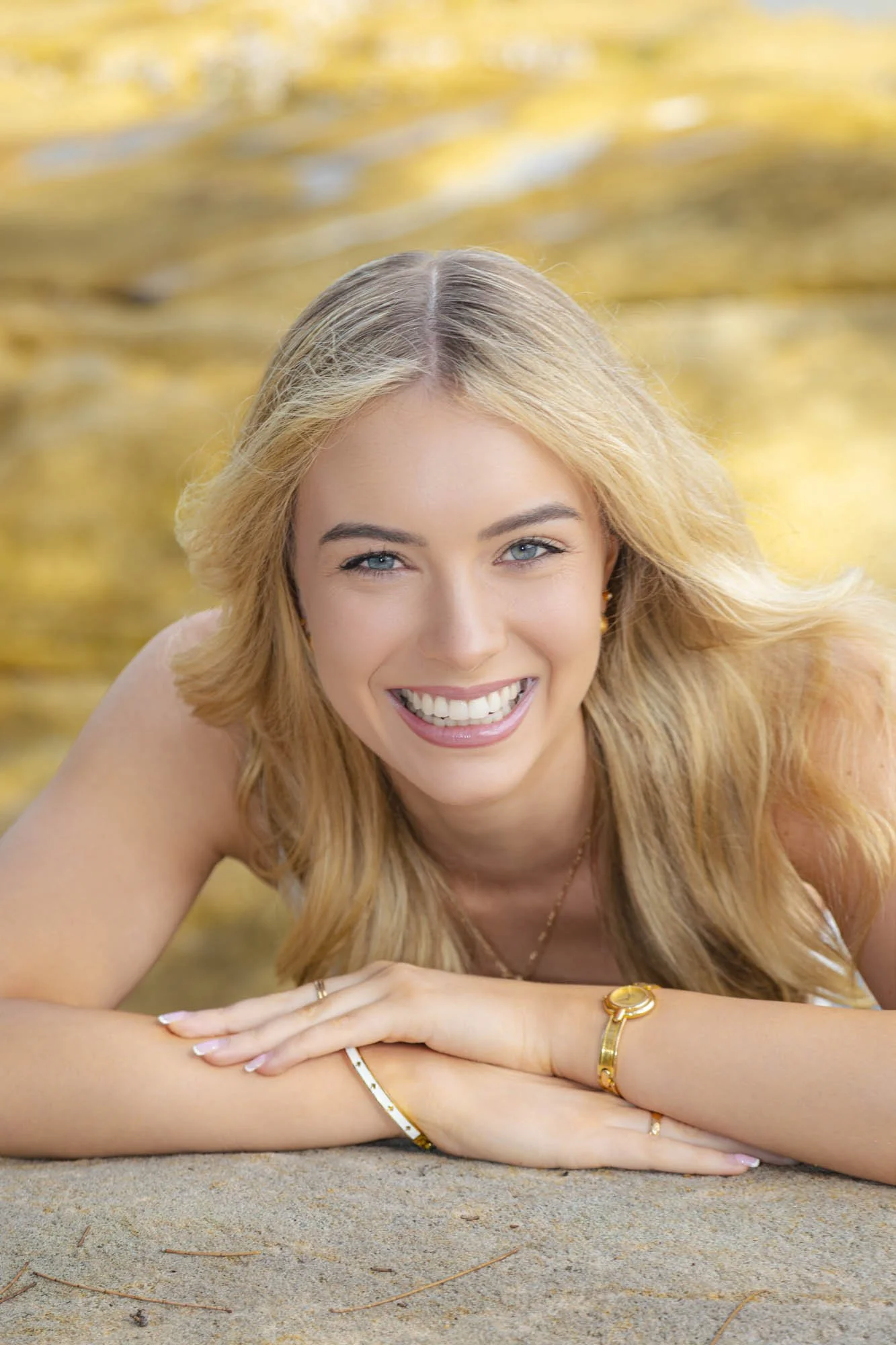 A woman with blonde hair smiling and lying on her stomach on a sandy surface with yellow autumn leaves in the background.