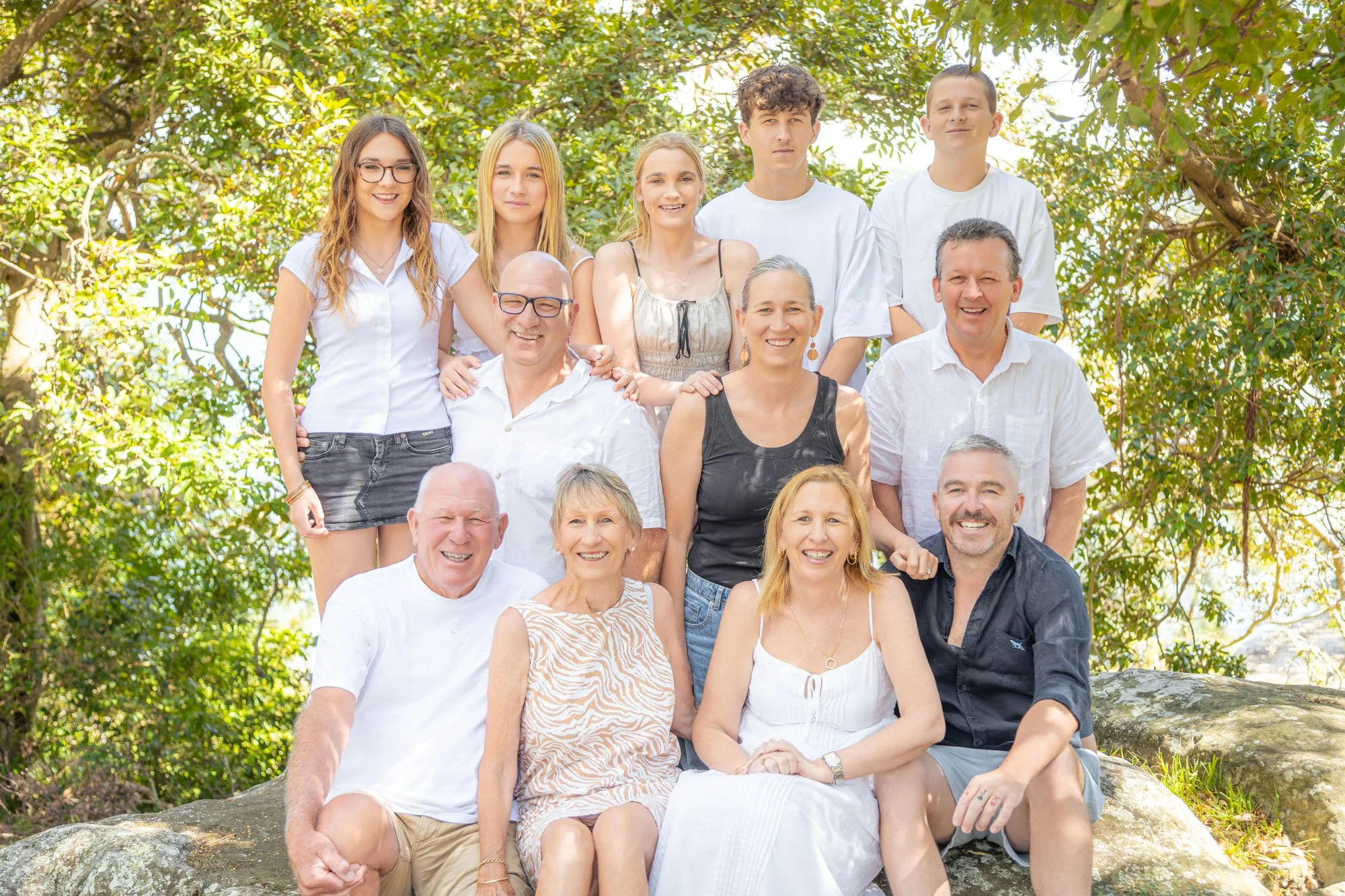 A multigenerational family photo outdoors with trees and sunlight, showing four older adults sitting on rocks in the front, three more older adults kneeling on rocks and five teenagers or young adults standing behind them, all smiling, during a relax