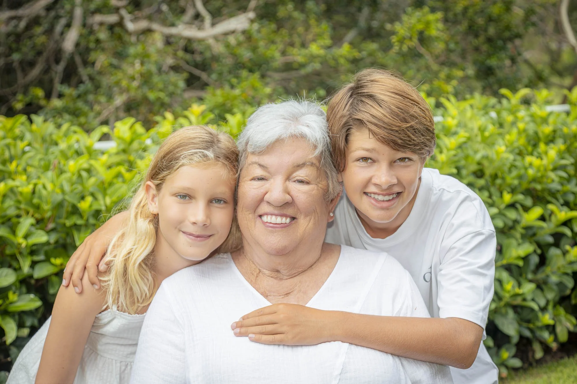 A grandmother with her two grandchildren, a girl and a boy, outdoors in a garden, smiling and hugging.