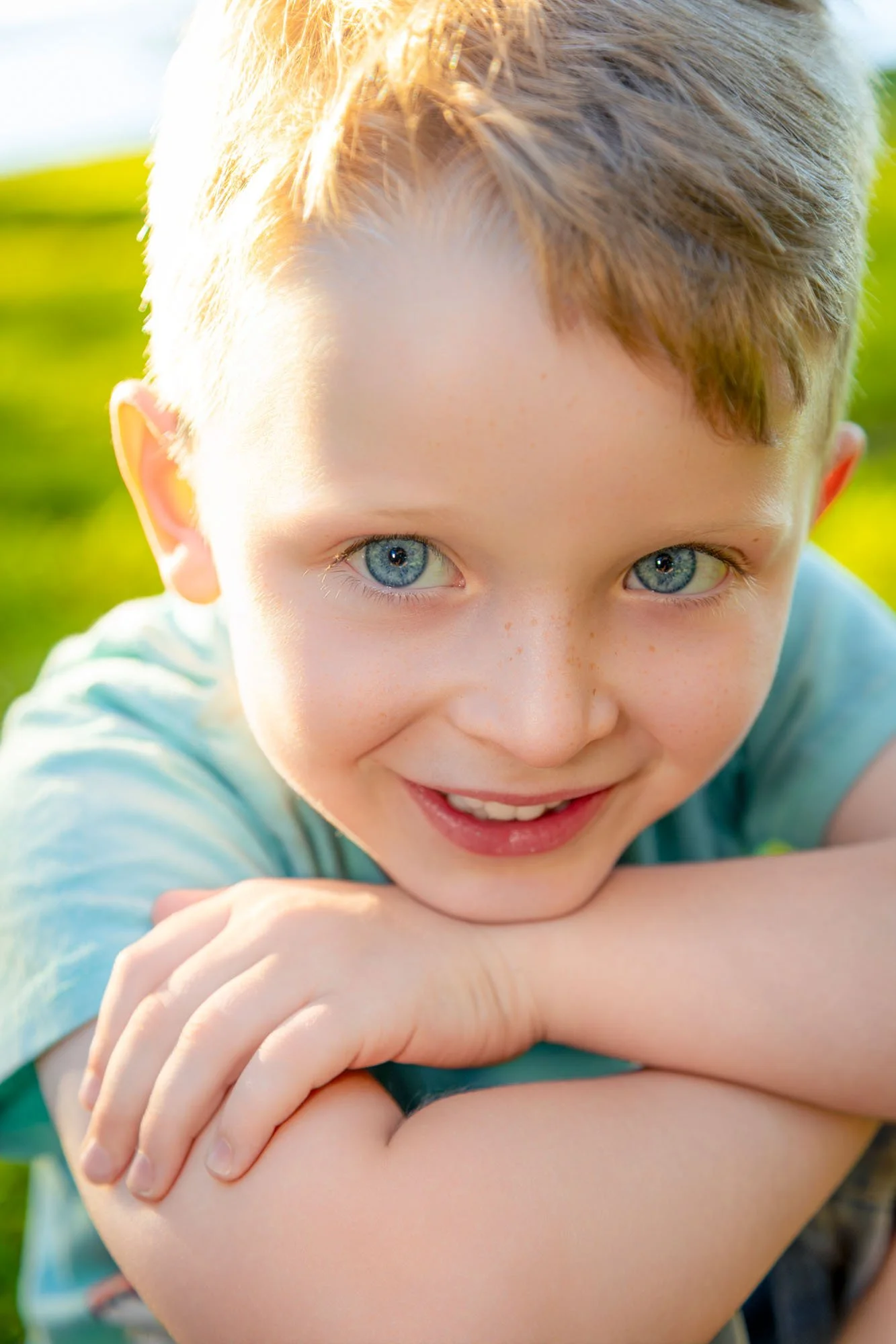 A Close-up of a smiling young boy with blue eyes and short light brown hair, resting his chin on his crossed arms outdoors in bright sunlight during a relaxed outdoor family photoshoot by Southern Exposure Photography.