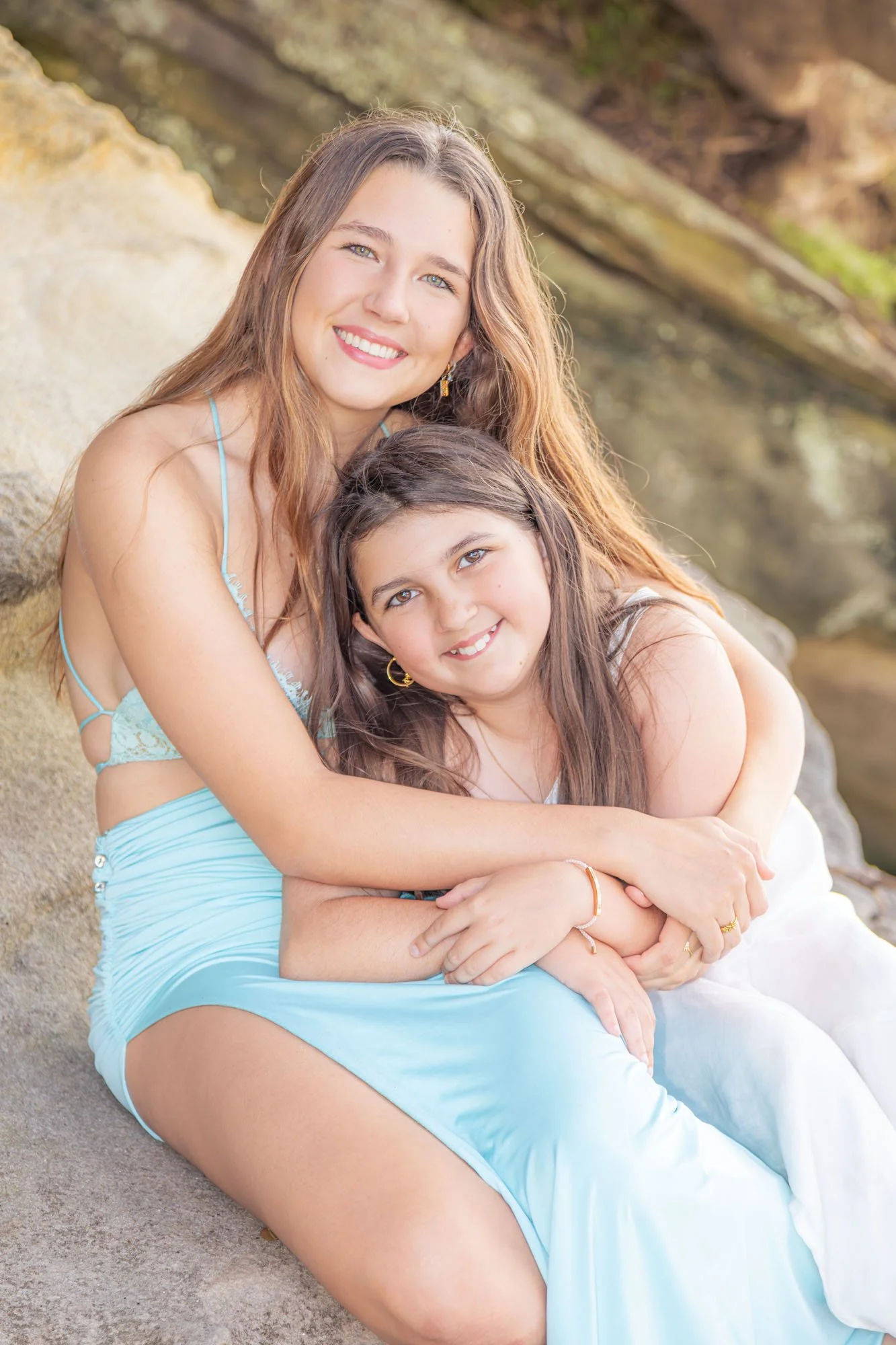 A group of two young sisters sitting on a rocky shoreline, embracing each other and smiling at the camera, in front of water and rocks during a relaxed outdoor family photoshoot by Southern Exposure Photography.