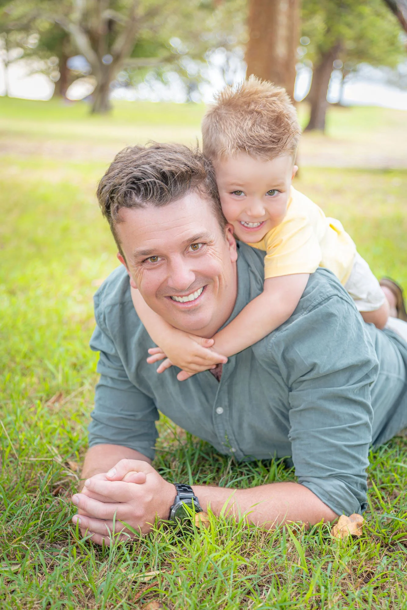 A man and a young boy lying on grass, with the boy on the man's back, both smiling at the camera in a park setting during a relaxed outdoor family photoshoot by Southern Exposure Photography.