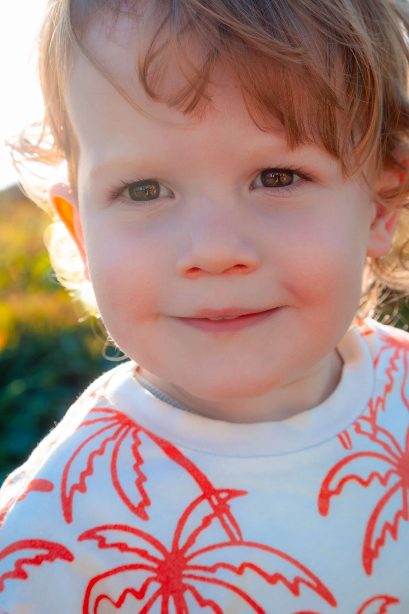 A close-up of a smiling young boy with curly red hair, wearing a white shirt with a red floral pattern, outdoors during golden hour during a relaxed outdoor family photoshoot by Southern Exposure Photography.