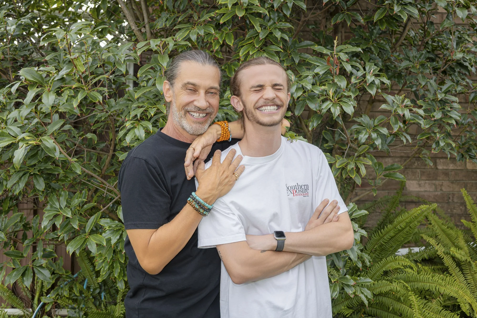 Two men smiling and laughing together outdoors, one with long gray hair and beard wearing a black shirt, the other with long light brown hair wearing a white t-shirt, with green foliage in the background.