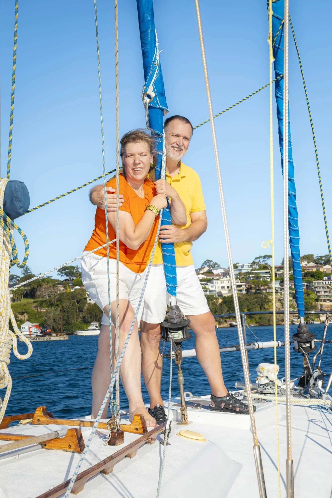 A woman and a man stand on a sailboat, smiling, with the woman holding the sailboat's mast and the man holding her waist, on a bright sunny day.