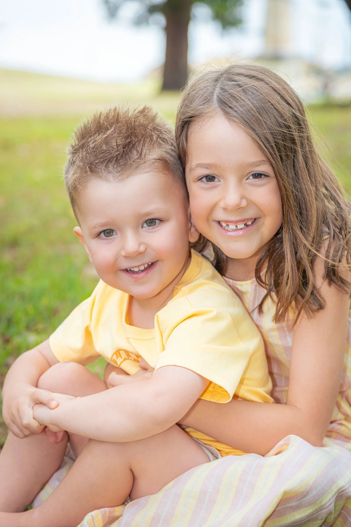 A group of two children, a brother and a sister, sitting outside and smiling at the camera during a relaxed outdoor family photoshoot by Southern Exposure Photography.
