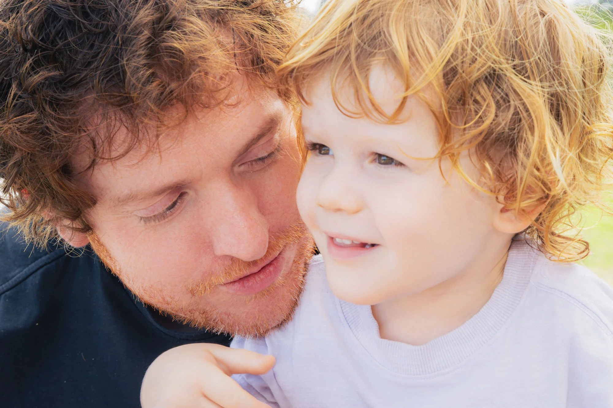 A father and a young son with curly red hair are close together outdoors, smiling and sharing a tender moment during a relaxed outdoor family photoshoot by Southern Exposure Photography.