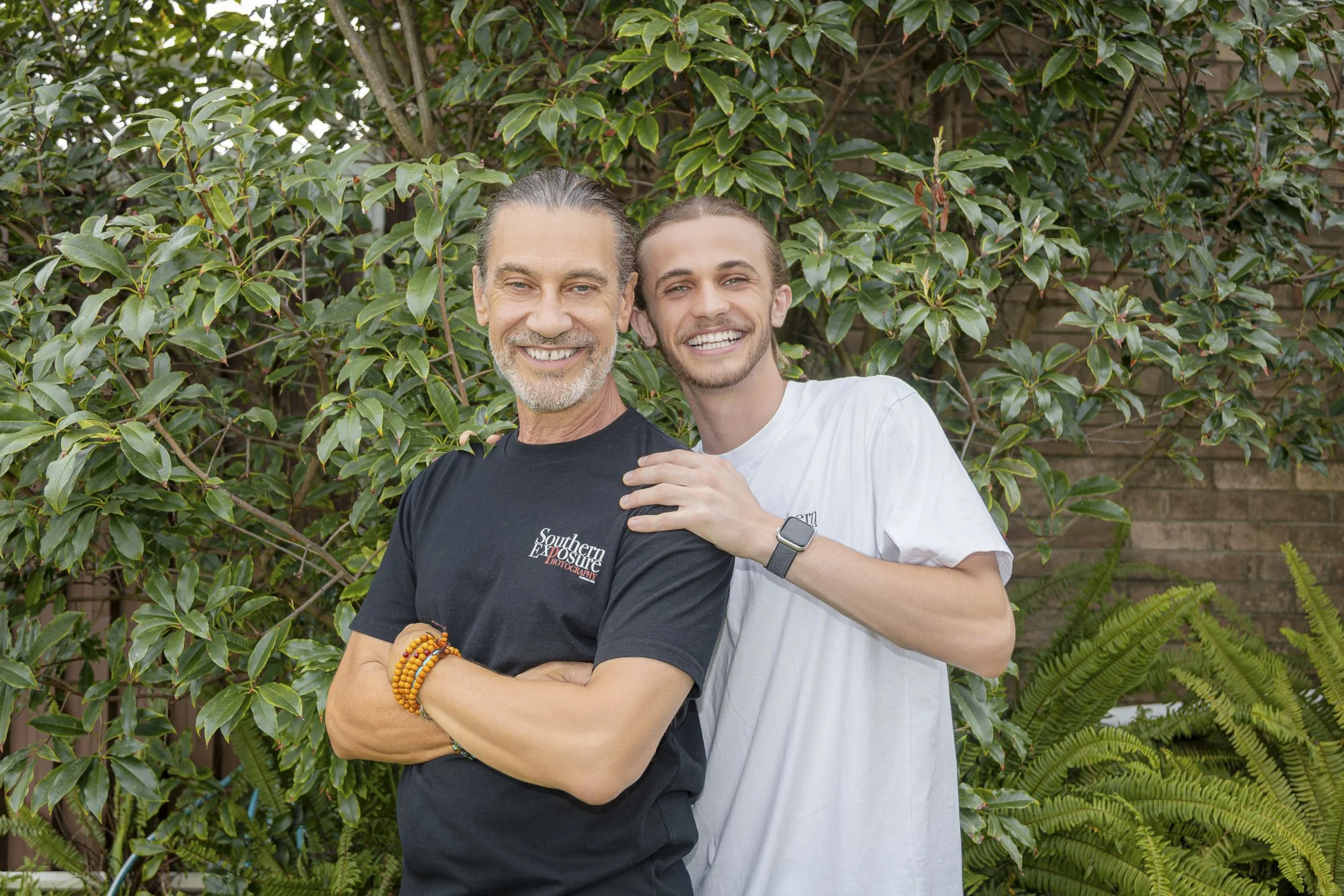 Two smiling men standing outdoors in front of green foliage, one with gray hair and a beard wearing a black t-shirt, the other with brown hair in a ponytail wearing a white t-shirt.