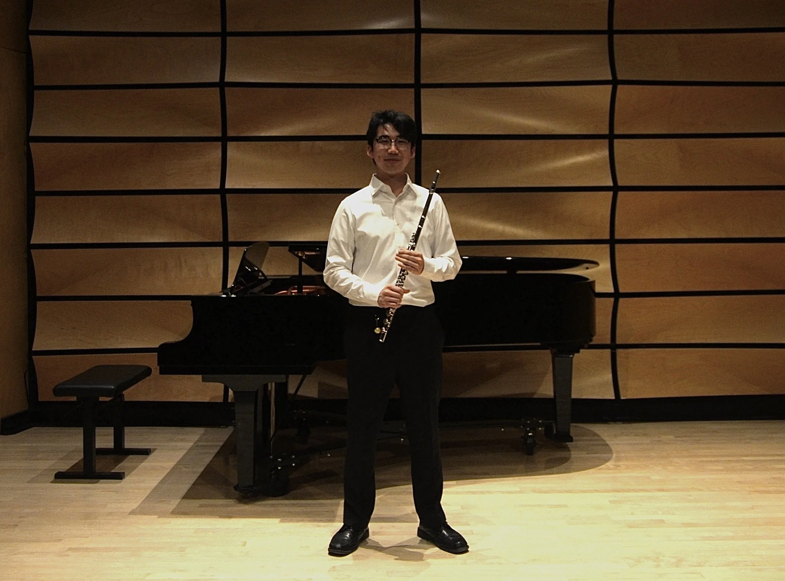 A man standing in front of a wooden wall with black horizontal and vertical lines, holding a flute, wearing glasses, a white shirt, black pants, and black shoes, with a grand piano behind him.