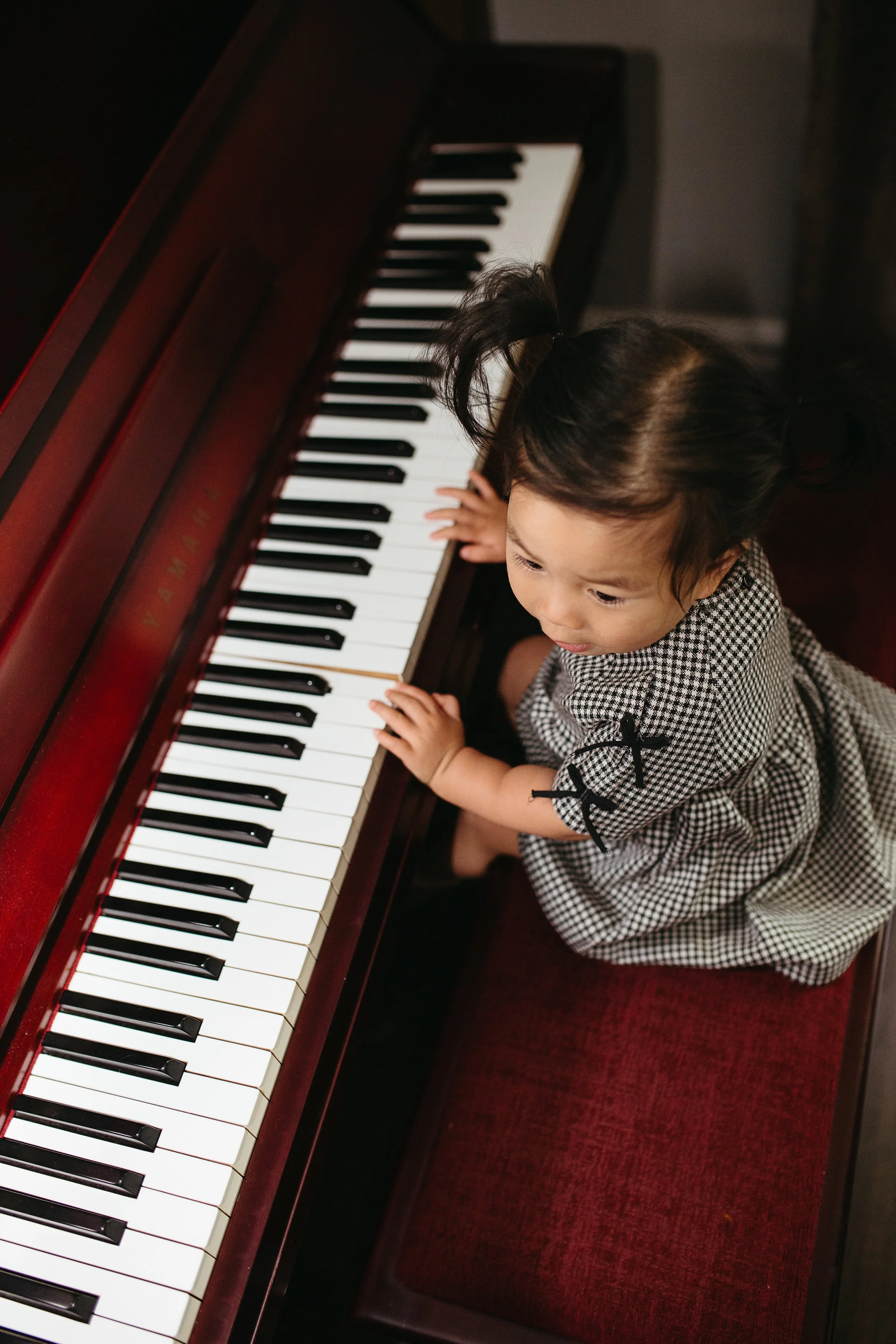 A young girl with dark hair in pigtails, wearing a checkered dress, sitting at a red upright piano and playing the keys.