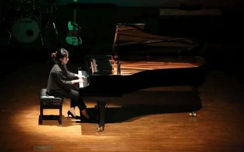 A woman playing grand piano on stage with musical instruments in background.