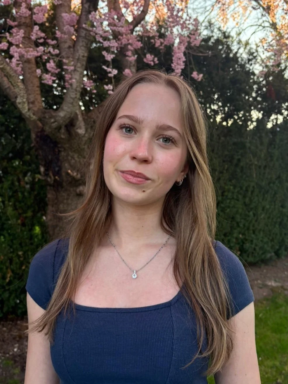 A young woman with light brown hair and blue eyes standing outdoors, with a pink flowering tree and green bushes in the background.