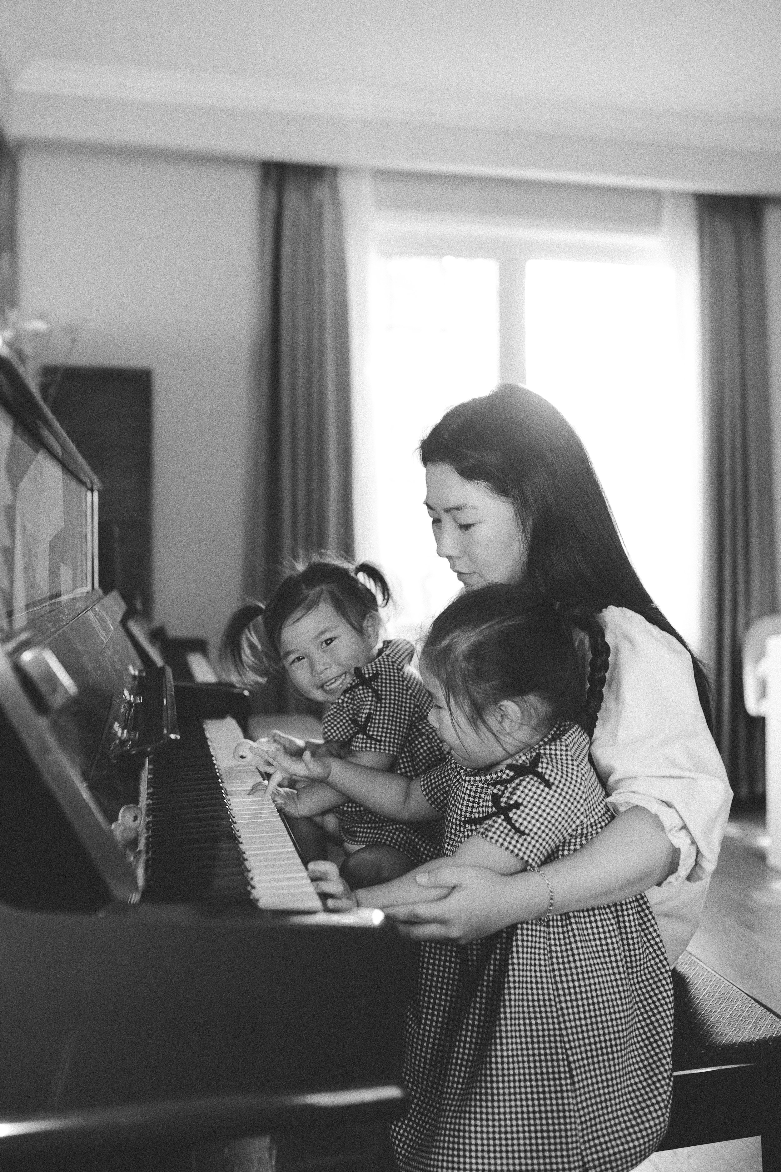 A woman helping two young girls play the piano together in a brightly lit room.