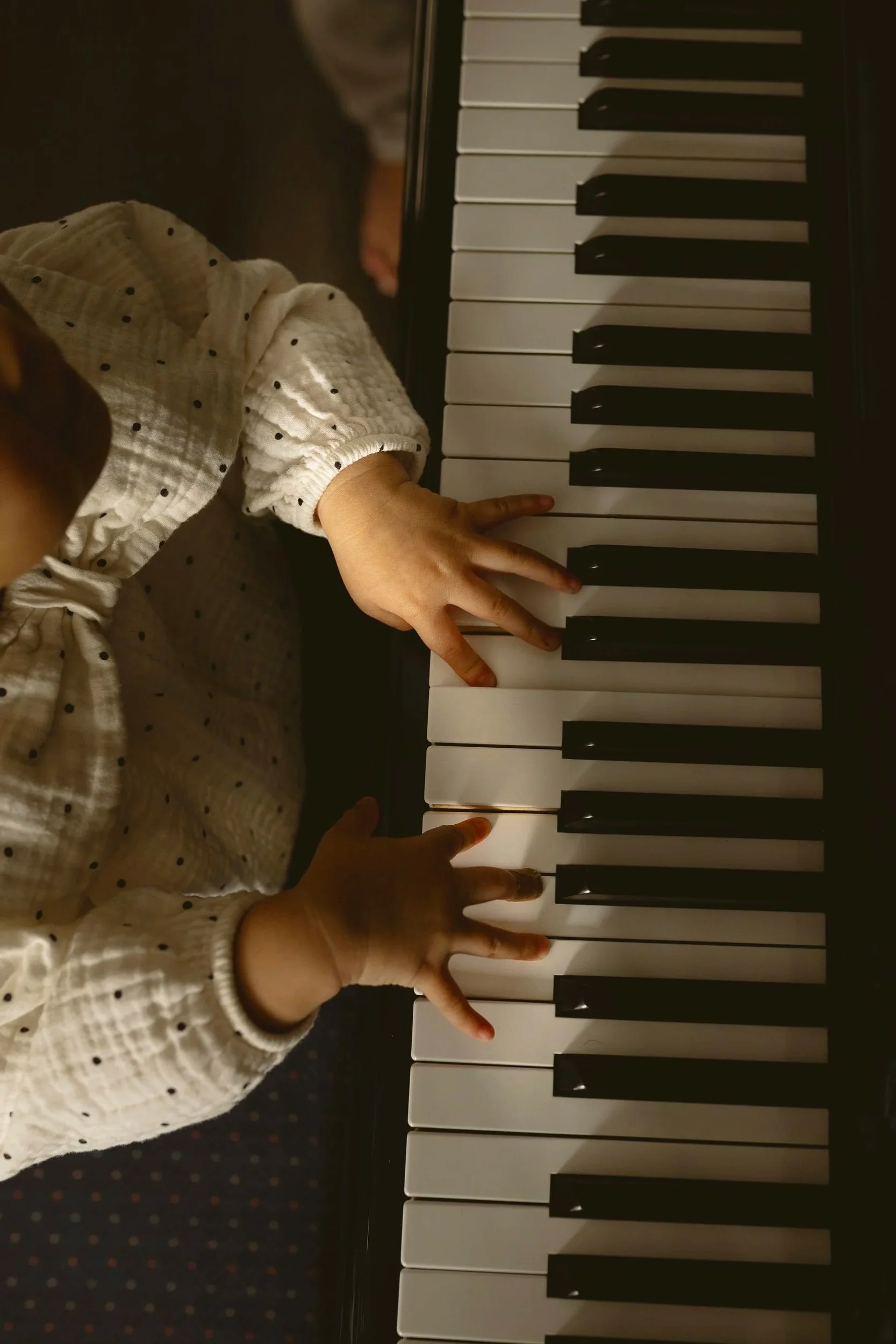 A child's small hands playing the piano, pressing black and white keys, wearing a long-sleeve shirt with black polka dots.