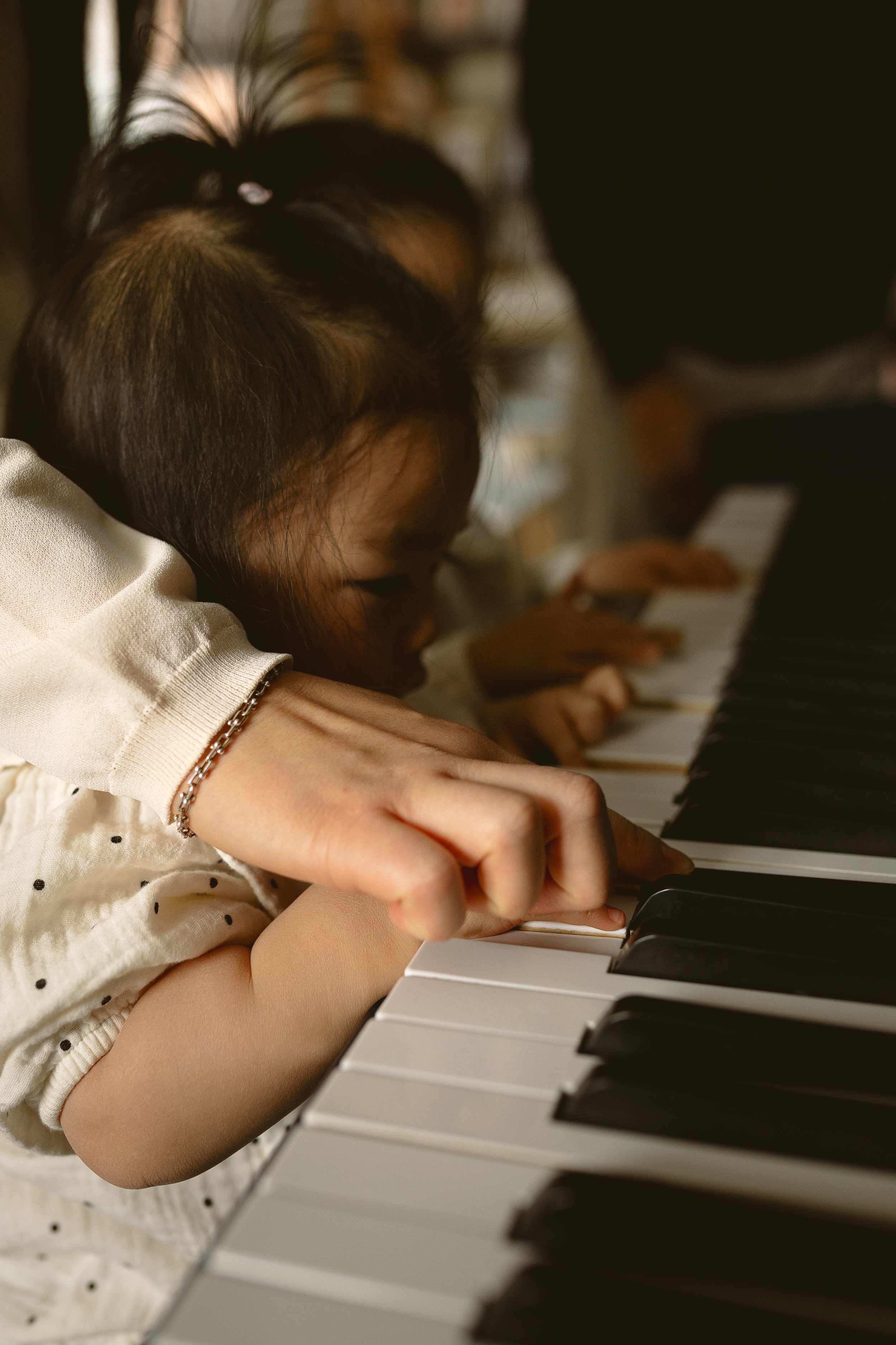 A young girl with dark hair is leaning over a piano, playing the keys with both hands. She is wearing a light-colored, polka-dotted shirt and a bracelet.