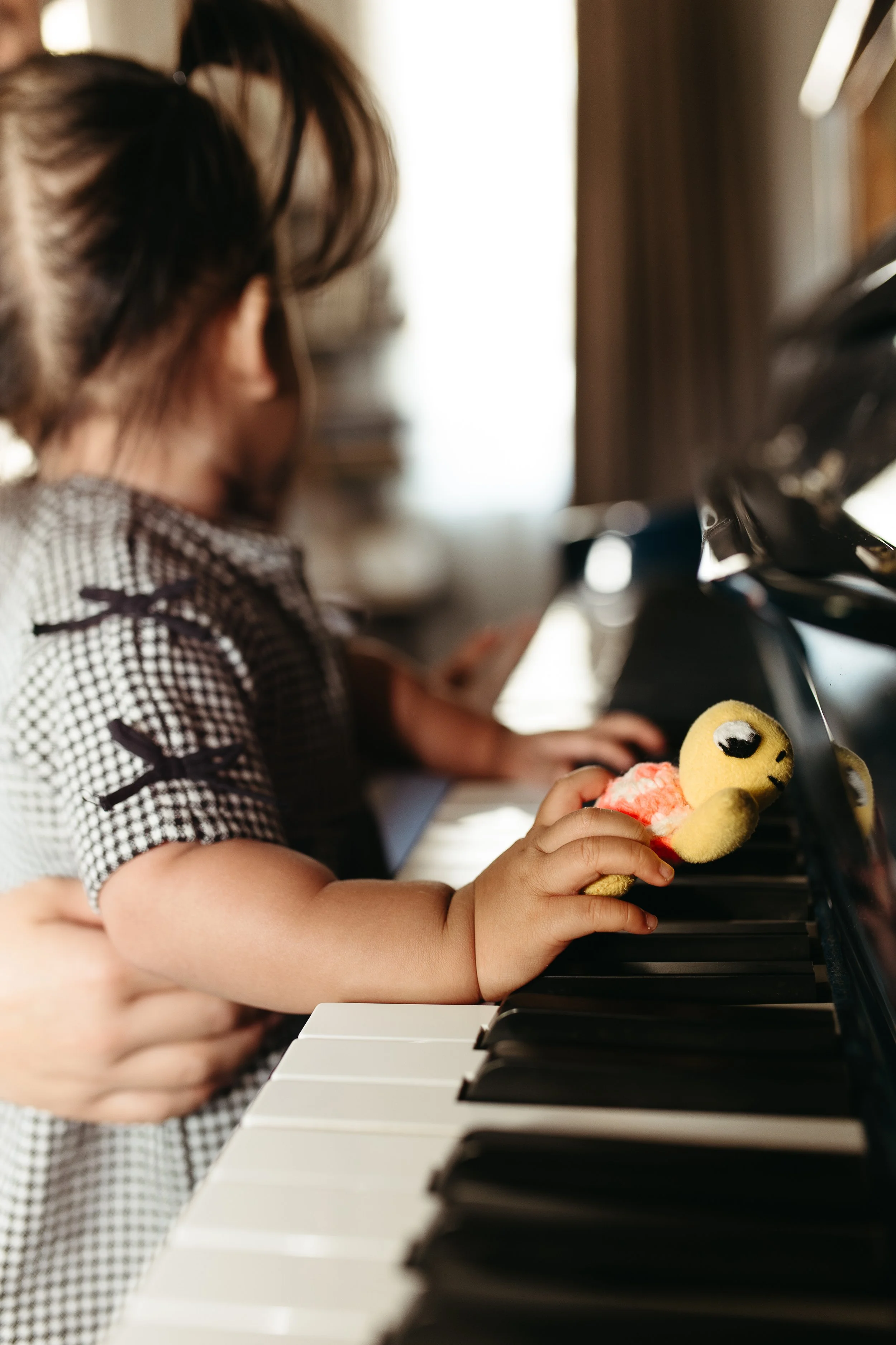 A young girl wearing a checkered shirt playing a black and white piano, holding a plush toy with a smiley face.
