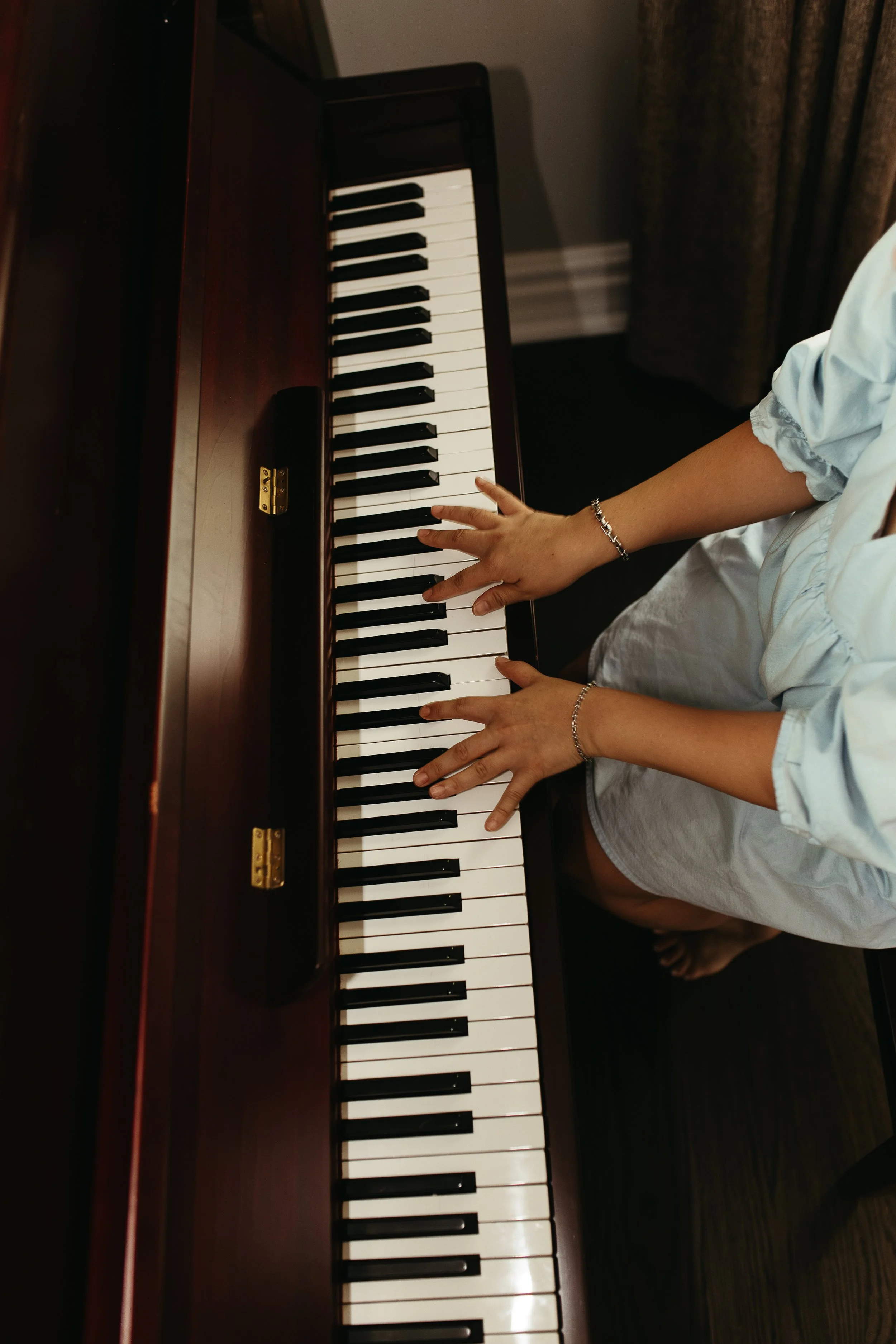 A person playing a wooden upright piano, dressed in a white shirt. The person's hands are on the piano keys, and they are seated on a dark floor near a wall with a curtain.
