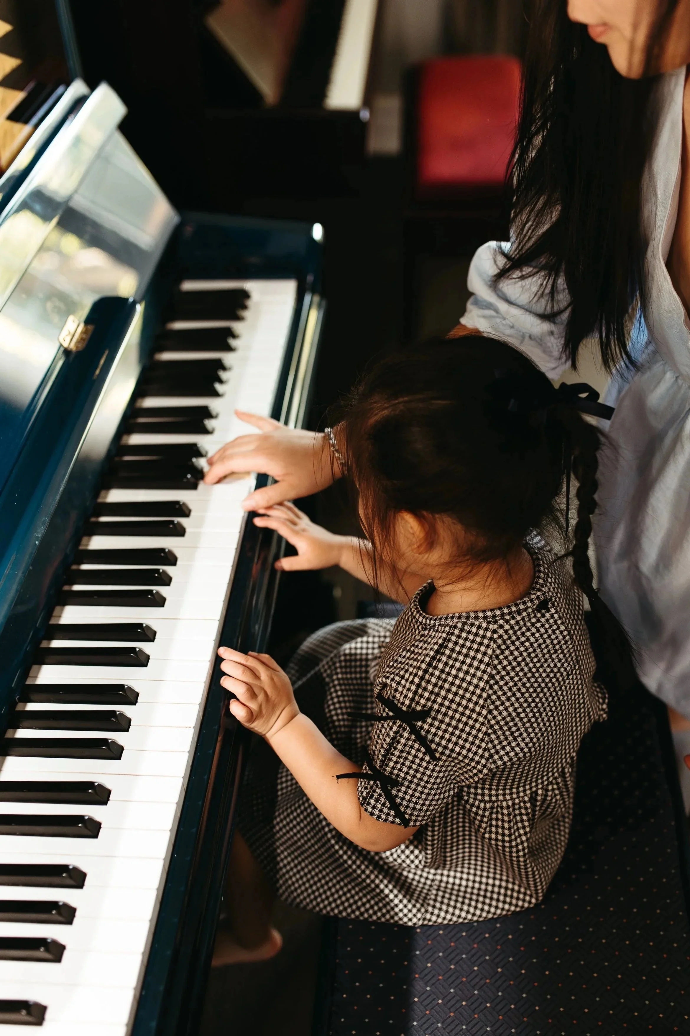 A young girl with dark hair in a braid playing a blue piano, seated on a dark patterned carpet in a room with a woman nearby.